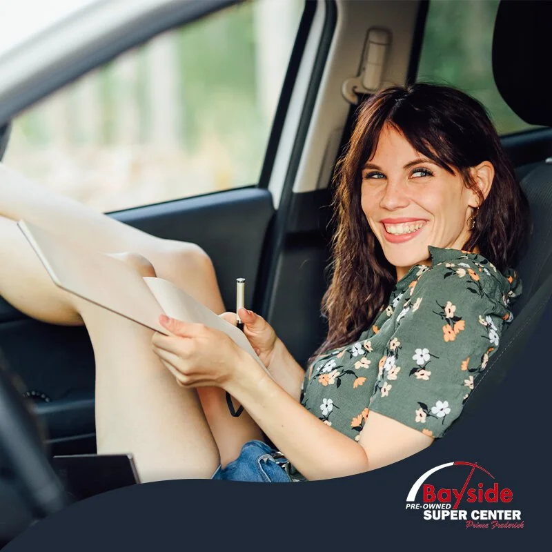 A woman sitting in a car, smiling, holding a pen and a notepad, wearing a floral shirt, with a blurred outside background.