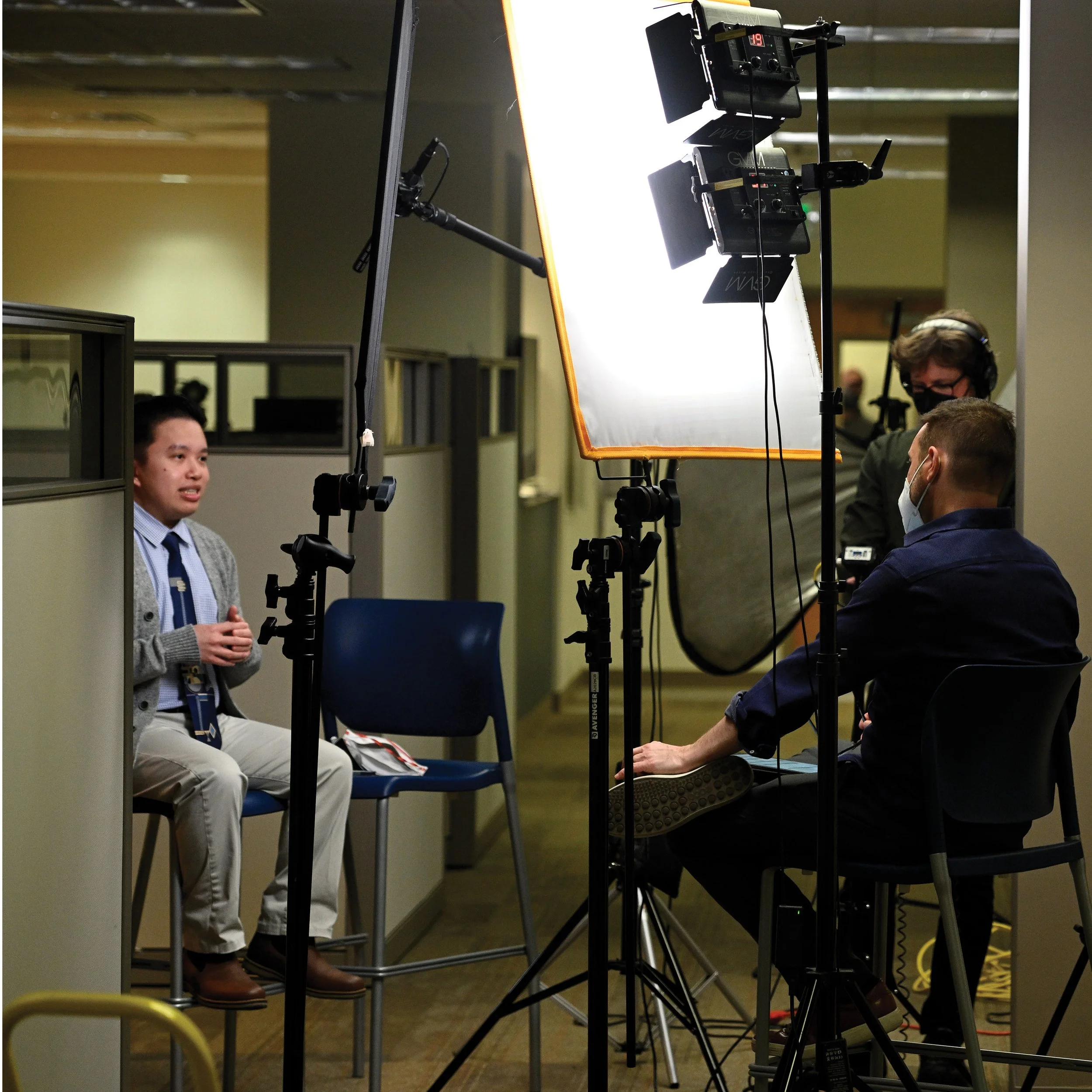 A young man in a gray cardigan and tie being filmed during an interview or recording session, with professional lighting and camera equipment set up in an indoor office environment.