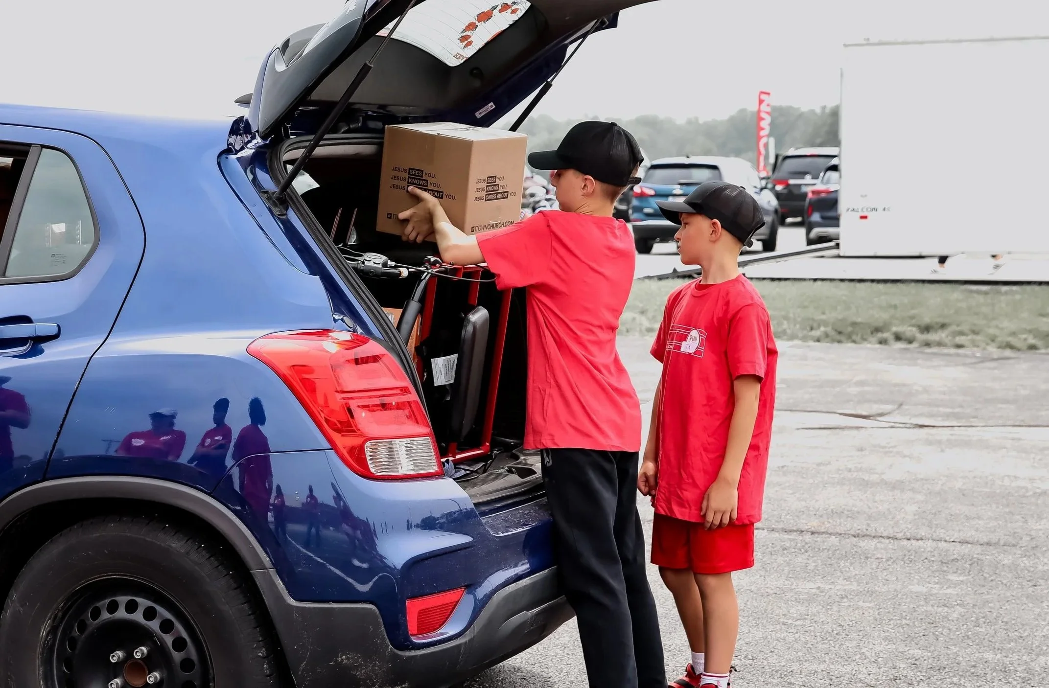 Two boys in red shirts and black caps loading a cardboard box into the trunk of a blue SUV at an outdoor parking lot.