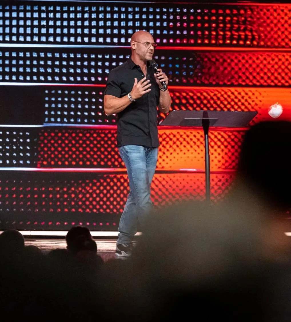 A man is speaking on stage, holding a microphone in his right hand, with a music stand in front of him, and a colorful, patterned background behind him.