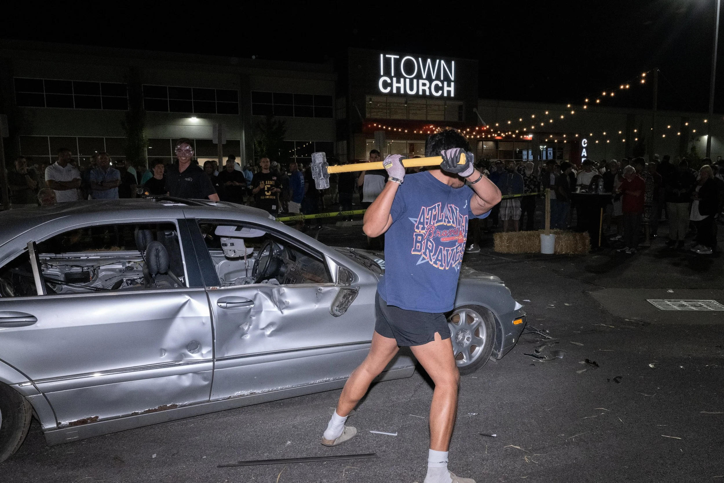 Nighttime scene outside a church with a large crowd. A person in shorts and a Braves t-shirt is holding a sledgehammer and standing near a damaged silver car with a crumpled door and dented side. The background features a lit-up sign reading 'ITOWN CHURCH' and string lights hanging overhead.