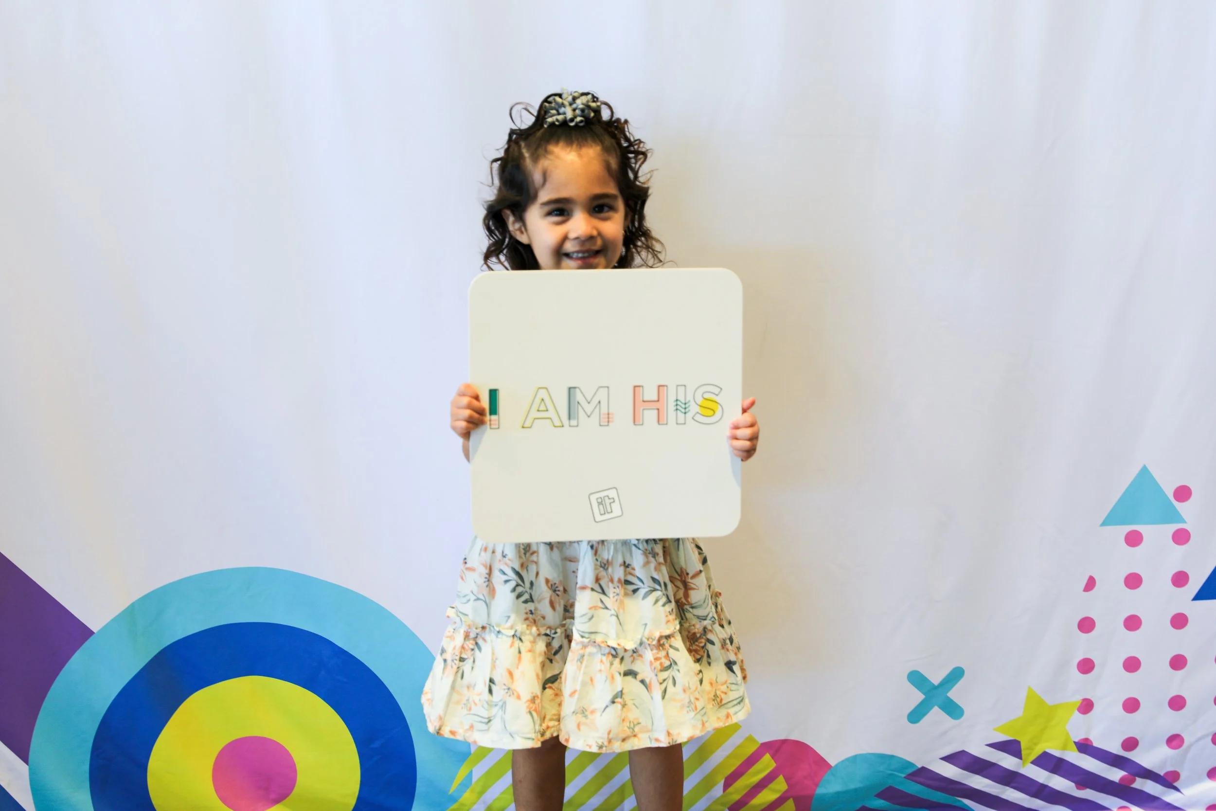 A young girl with curly hair and a bow, smiling and holding a white sign with colorful letters spelling 'I AM A HIS' in front of a colorful, playful background.