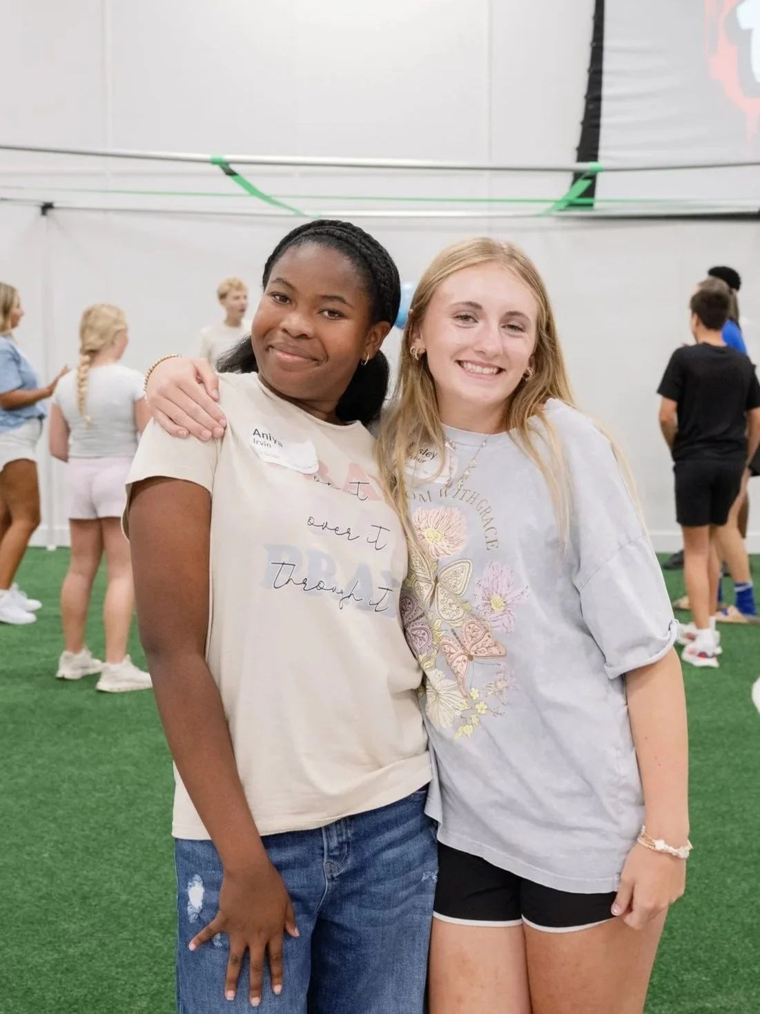 Two young women standing close together indoors on a green artificial turf. One has dark skin, black braided hair, wearing a beige t-shirt and blue jeans. The other has light skin, blond hair, wearing a gray graphic t-shirt and black shorts. They are smiling, with other people blurred in the background.