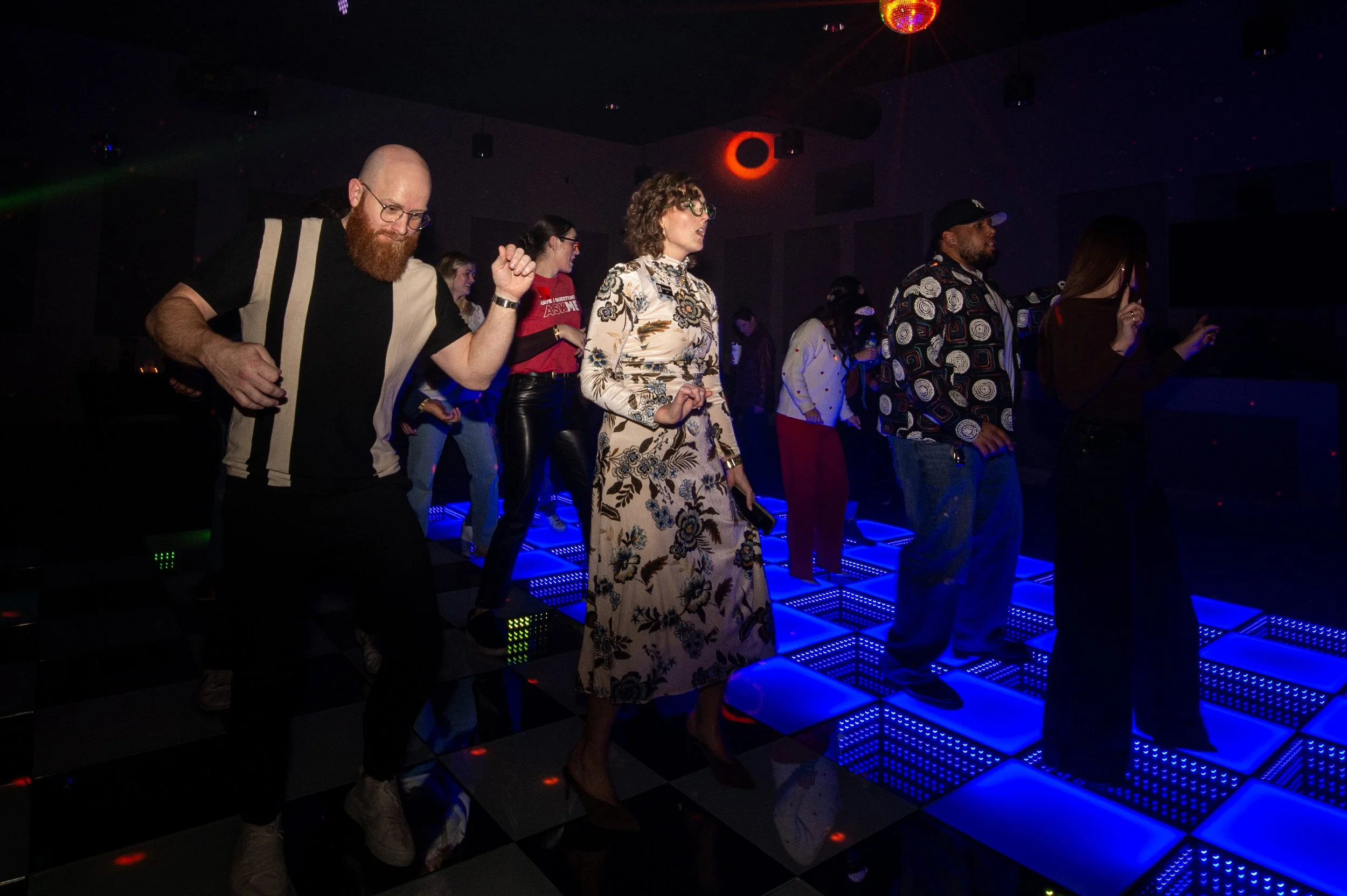 People dancing on a blue-lit dance floor in a dark room with a disco ball and colored lights.