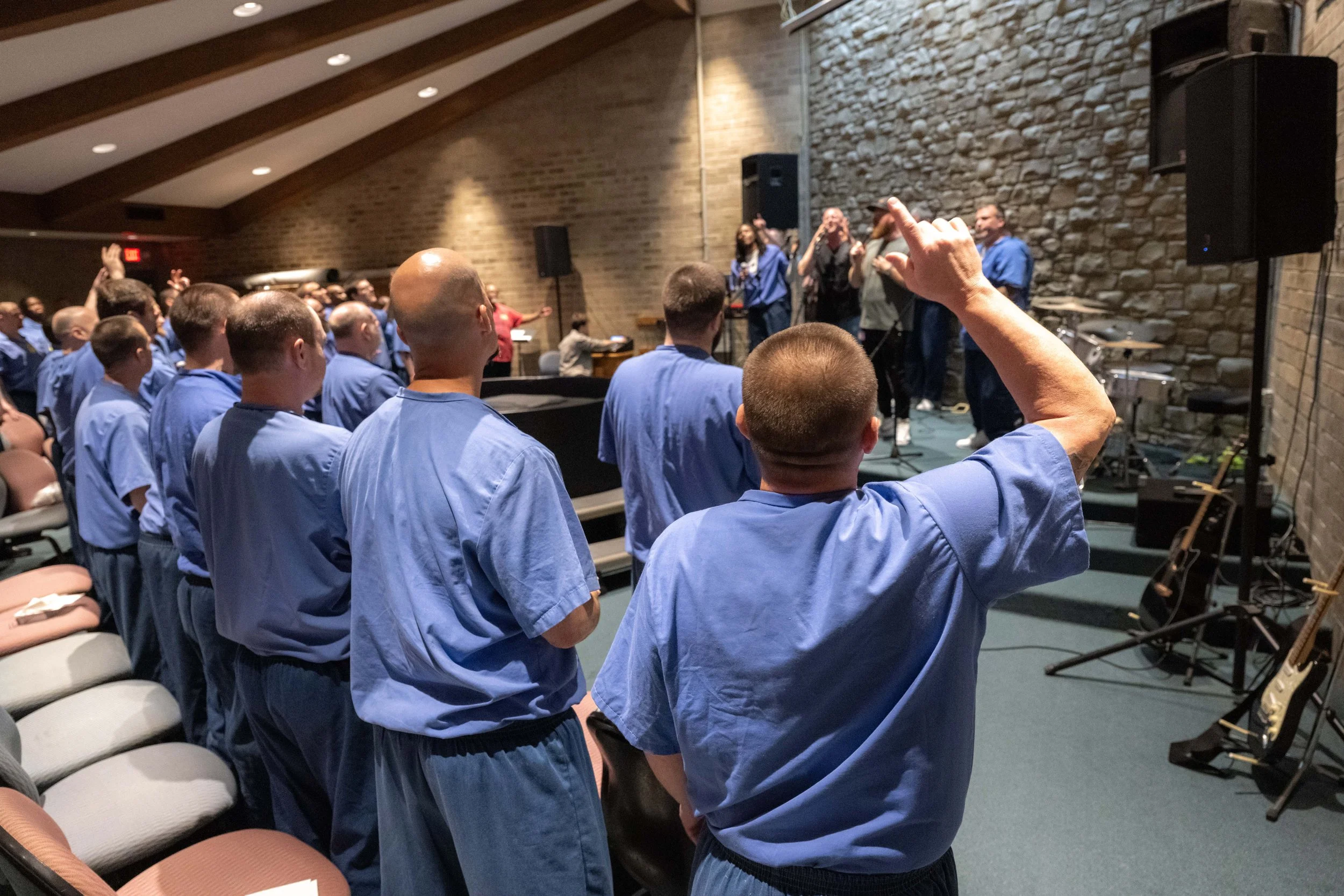Inmates standing in an auditorium with a stone wall and a band setup, participating in a worship service.