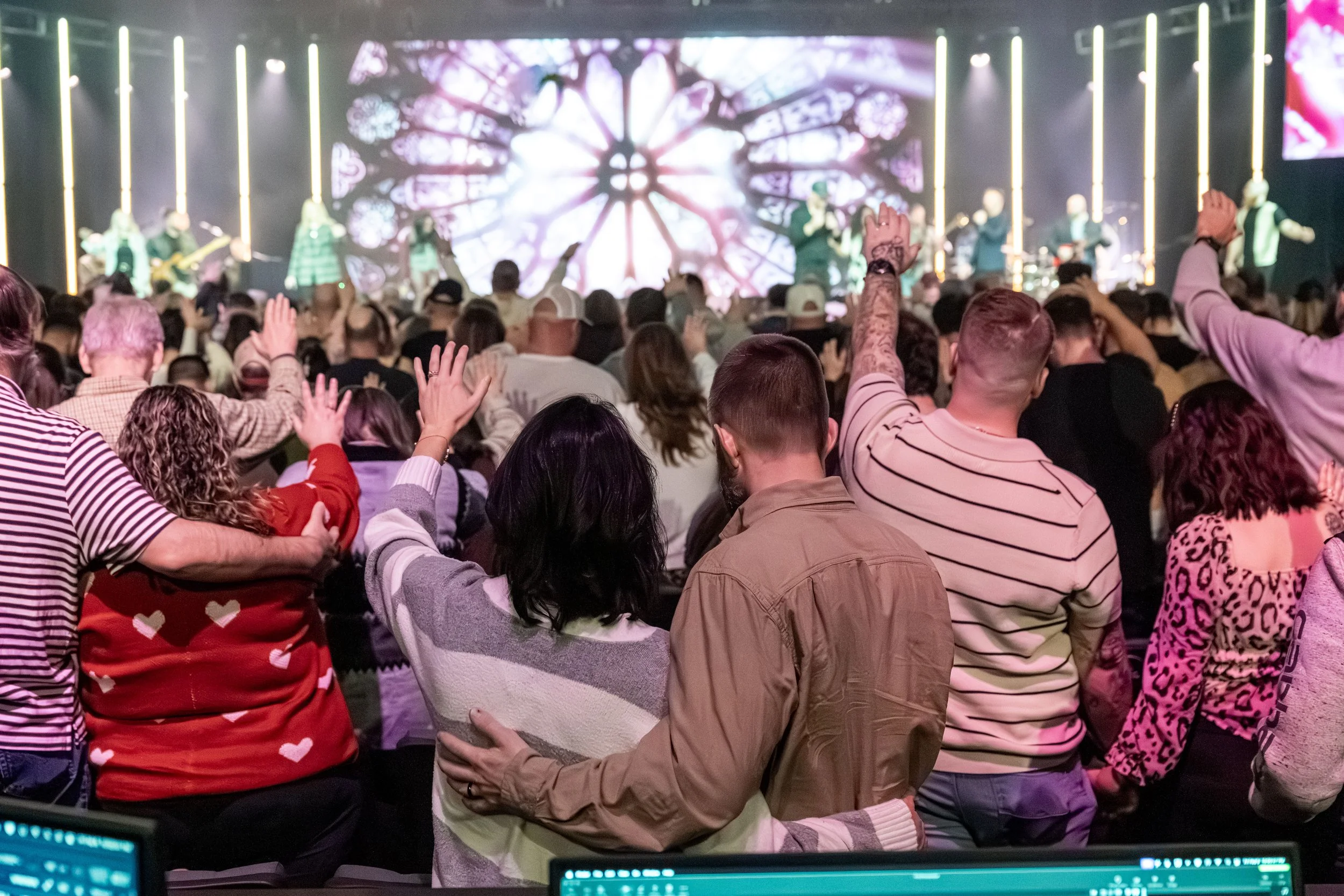 A large crowd of people at an event, raising their hands. In the background, there is a stage with a large digital screen displaying colorful abstract visuals.