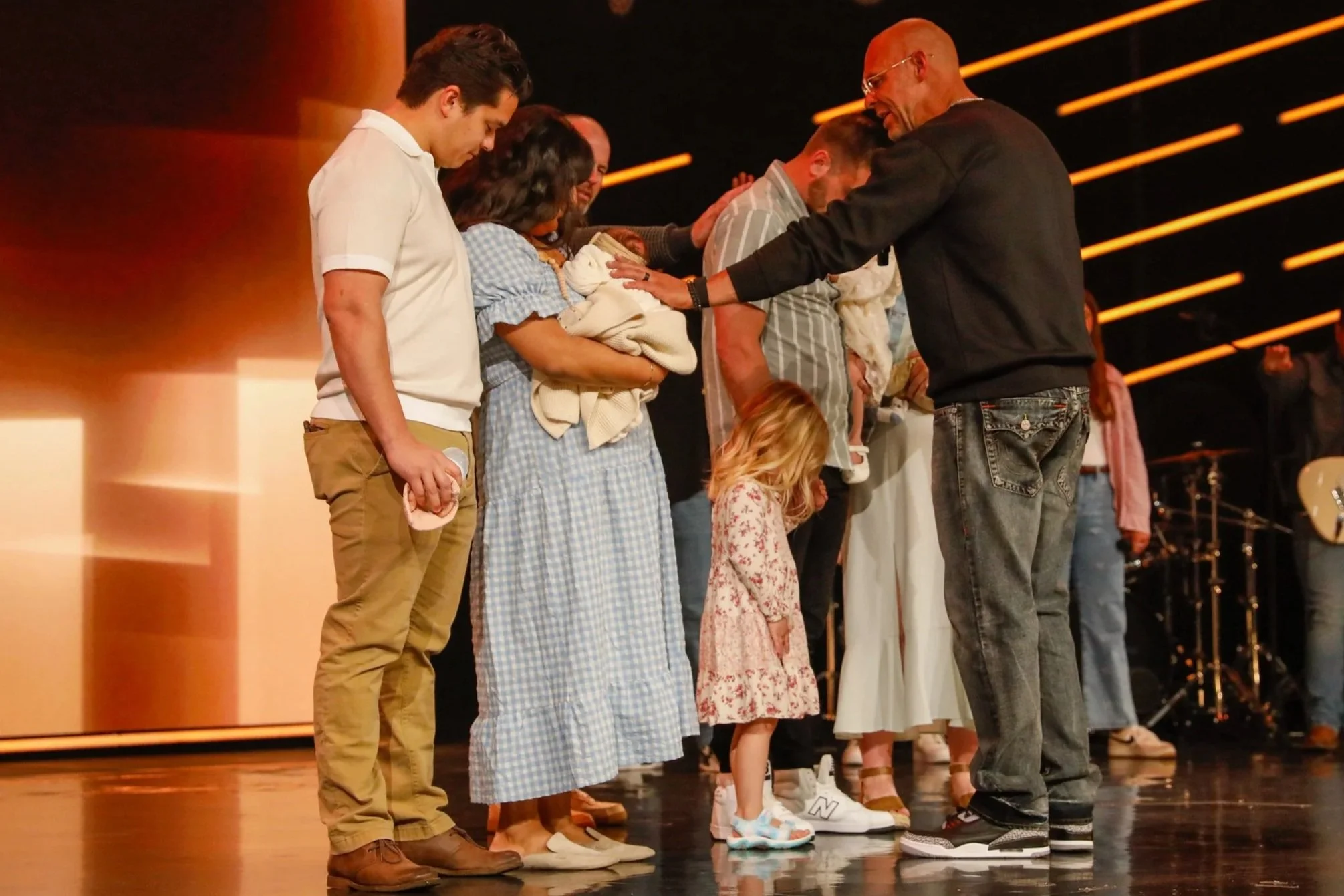 People standing in a row on stage, with their heads bowed and hands resting on a person's shoulders or head, during a church service blessing.