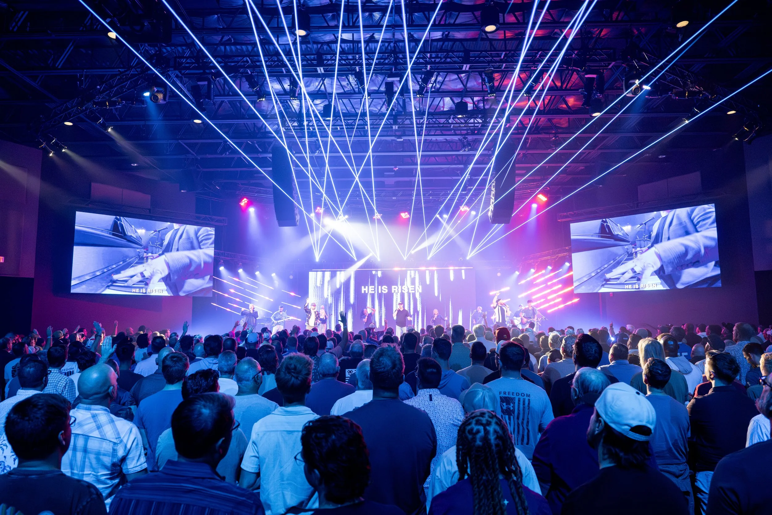 Crowd attending a church service with a large stage, musicians performing, and colorful lighting effects with laser beams, large screens displaying the event, and the phrase "HE IS RISEN".