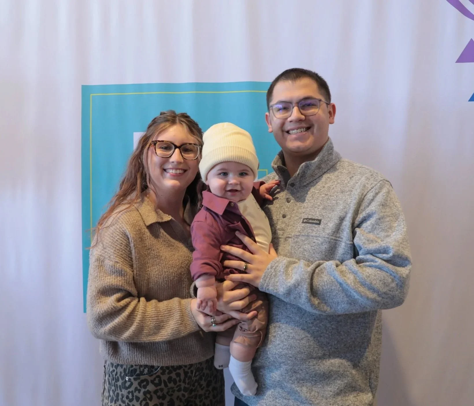 A family of three smiling and holding their young child in front of a light blue background with colorful design elements.