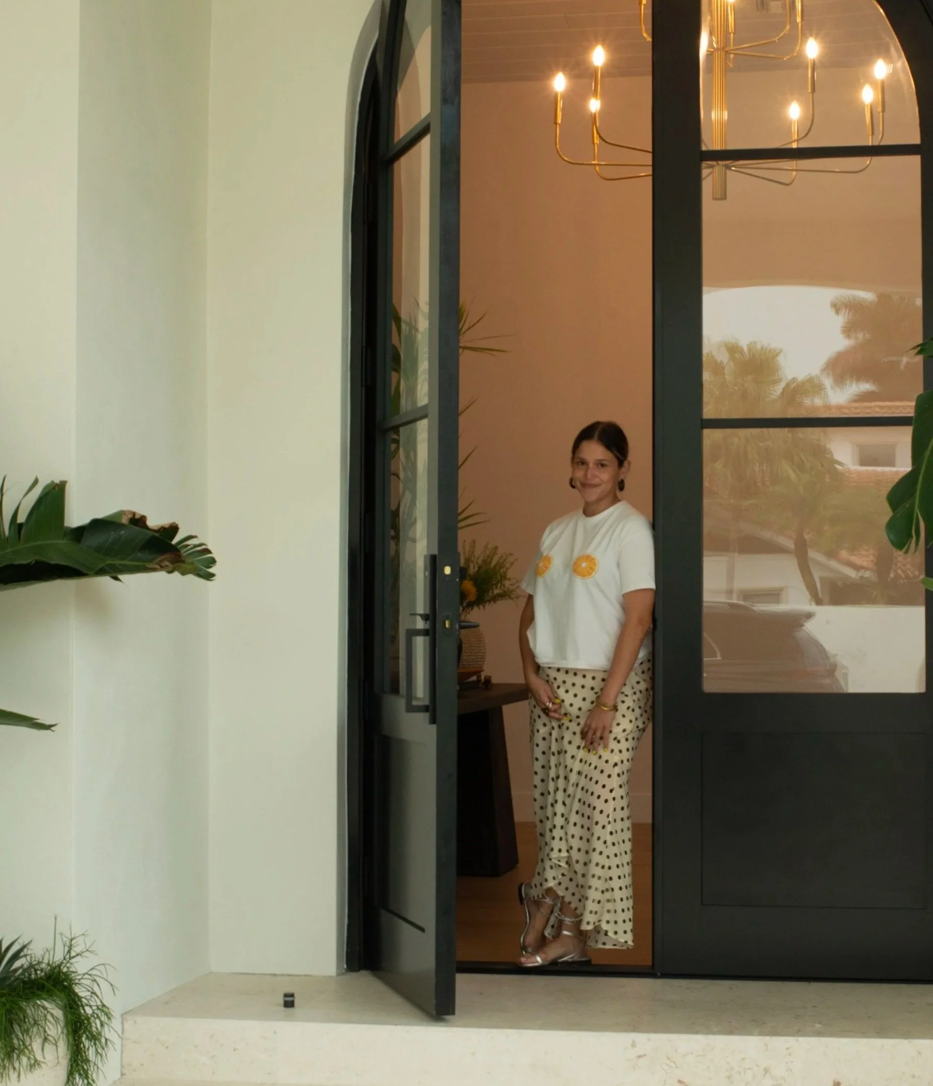 A woman standing inside a house, visible through an open black-framed glass door, smiling and wearing a white t-shirt with lemon prints, beige polka-dot pants, and open-toe heels. The background includes a chandelier, a houseplant, and reflections of trees outside.