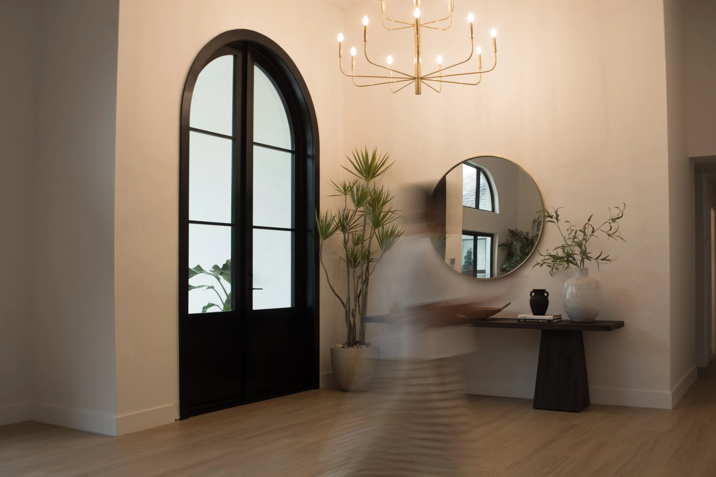 Interior of a modern home with a black arched window, gold chandelier, large mirror, potted plant, decorative vases, and a console table with books.