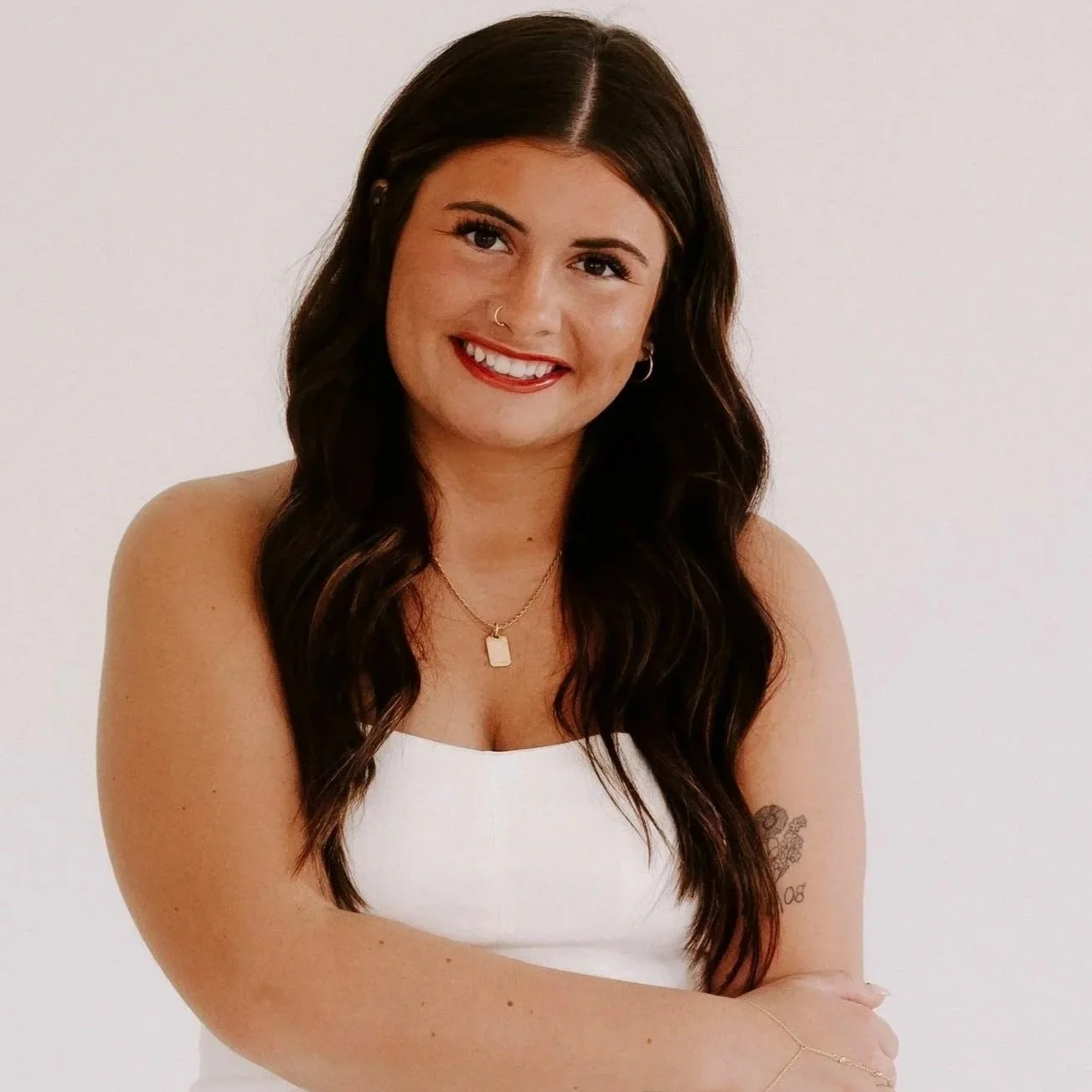 Smiling young woman with long dark hair, wearing a strapless beige top, a necklace, and bracelets, standing against a black background.