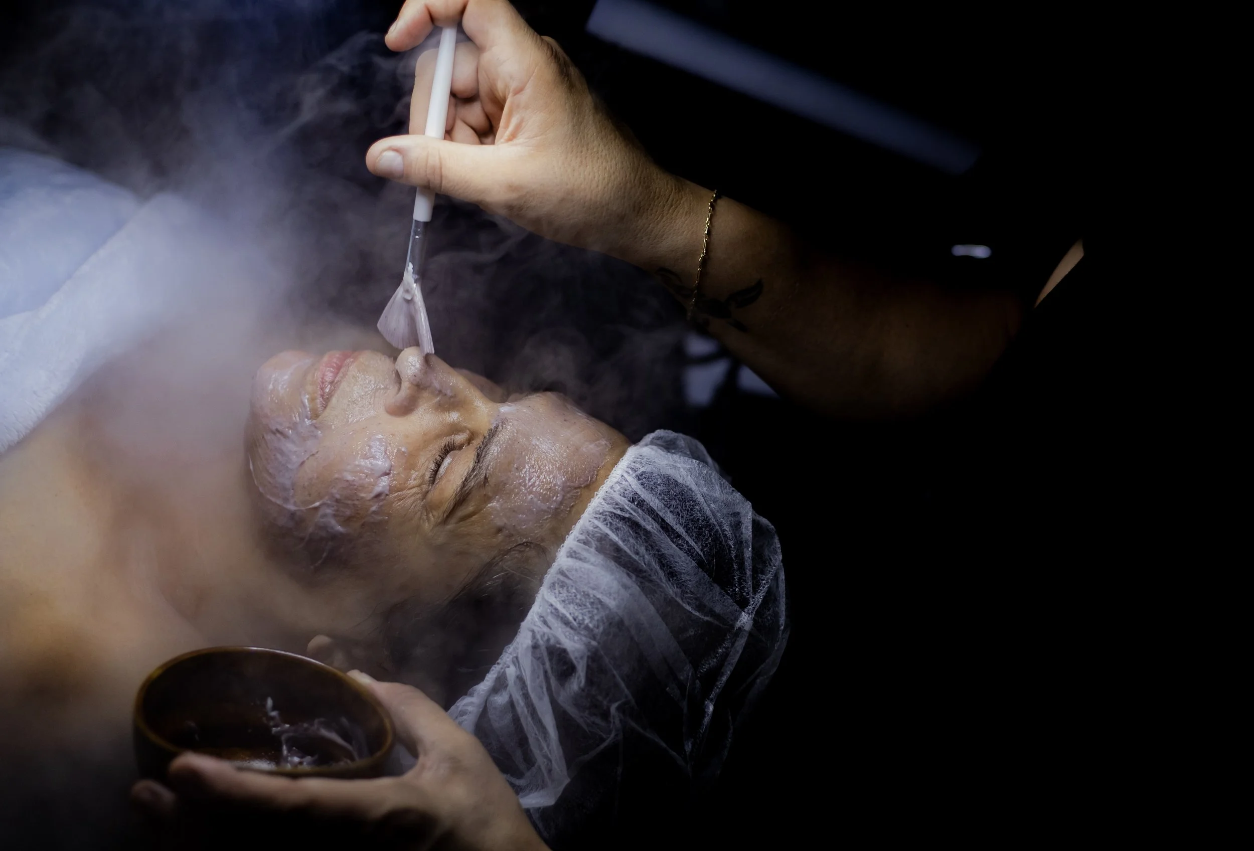 A person receives a facial treatment in a spa or clinic setting, with a beautician applying a mask or facial product, while steaming the face for skin rejuvenation.
