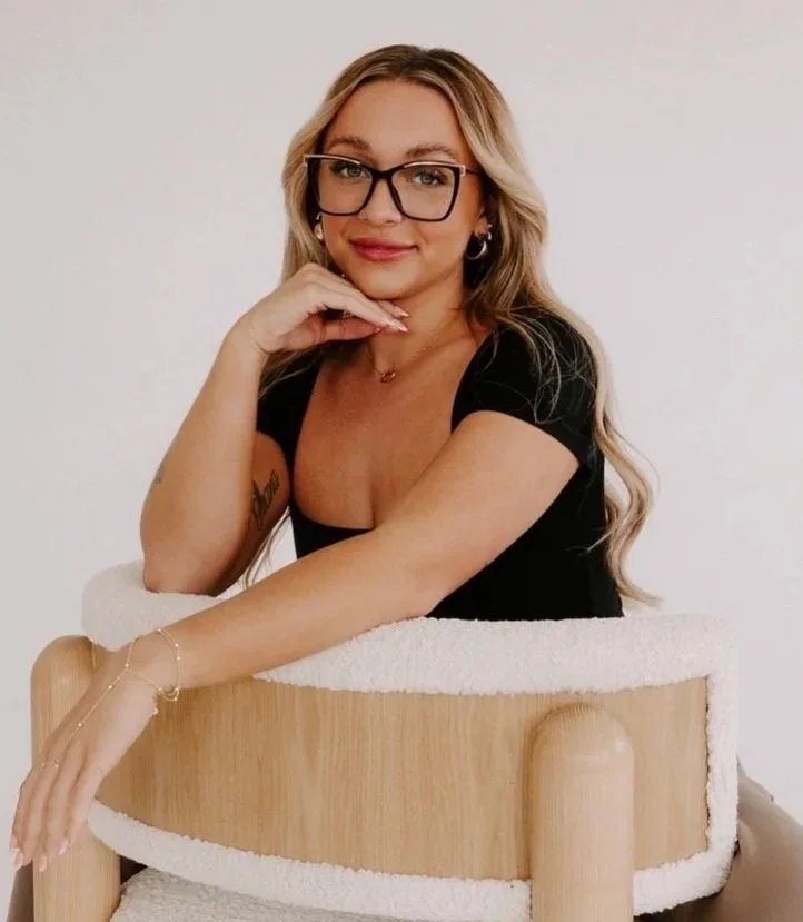 Woman with long, wavy blonde hair, wearing glasses, a black and white striped sleeveless dress, and tattoos on her arms, standing against a solid black background.