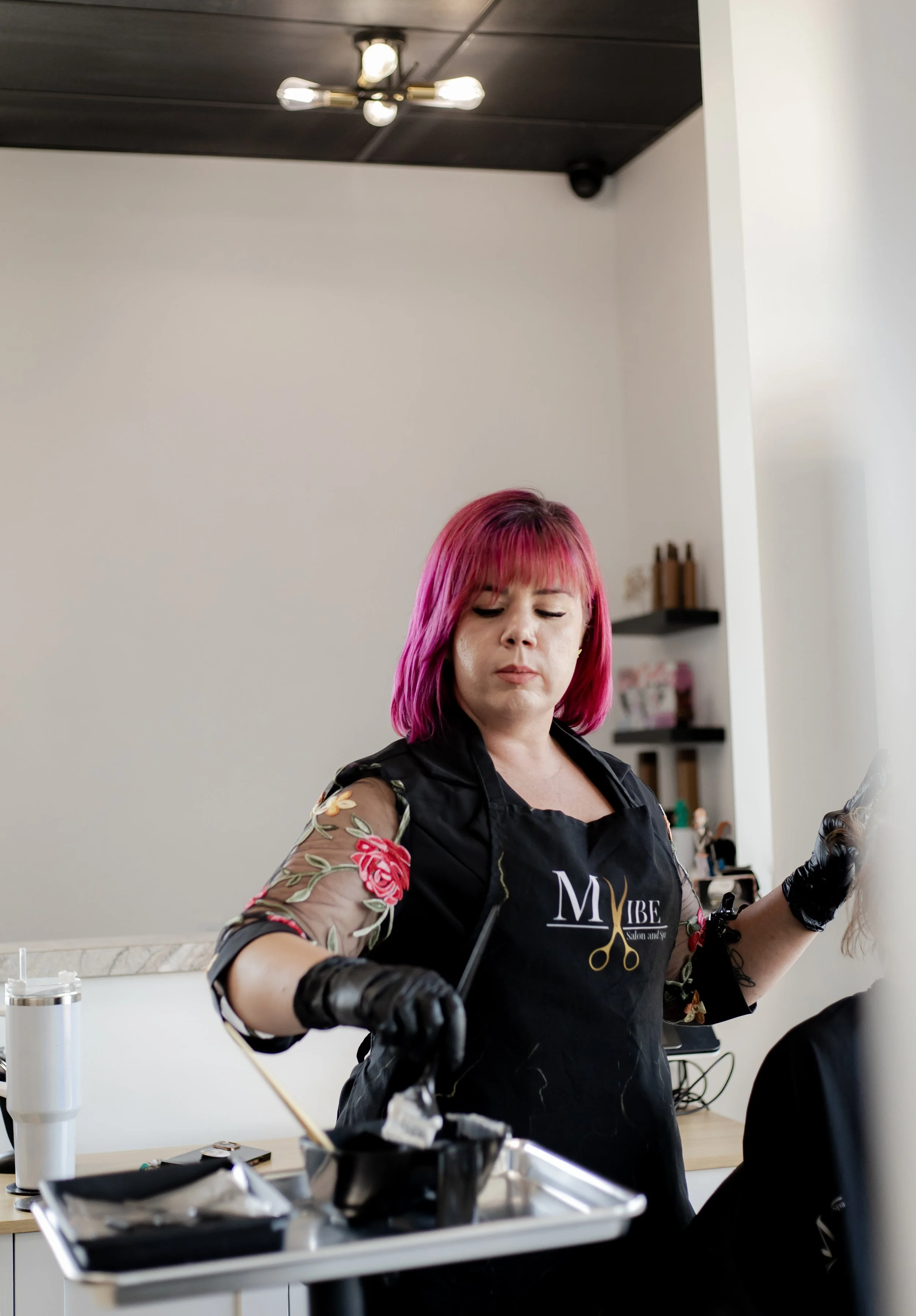 A woman with bright pink hair and a black apron working at a salon or barbershop, holding a tool and working on a client's hair.