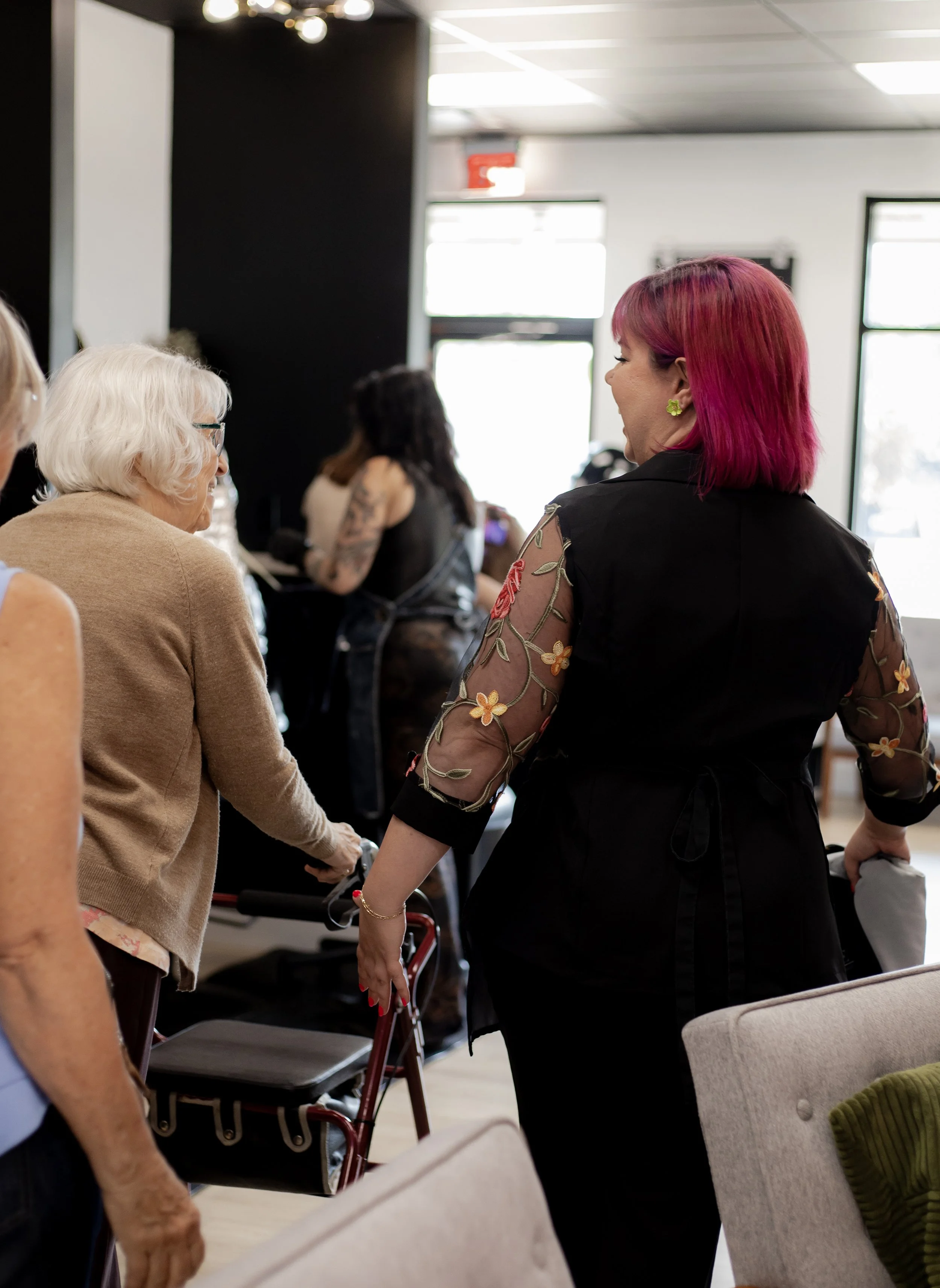 A group of women in a social setting, engaged in conversation. The woman on the right has short, pink hair and is wearing a black dress with sheer, floral embroidered sleeves. An older woman with white hair and glasses is using a walker, and other wo