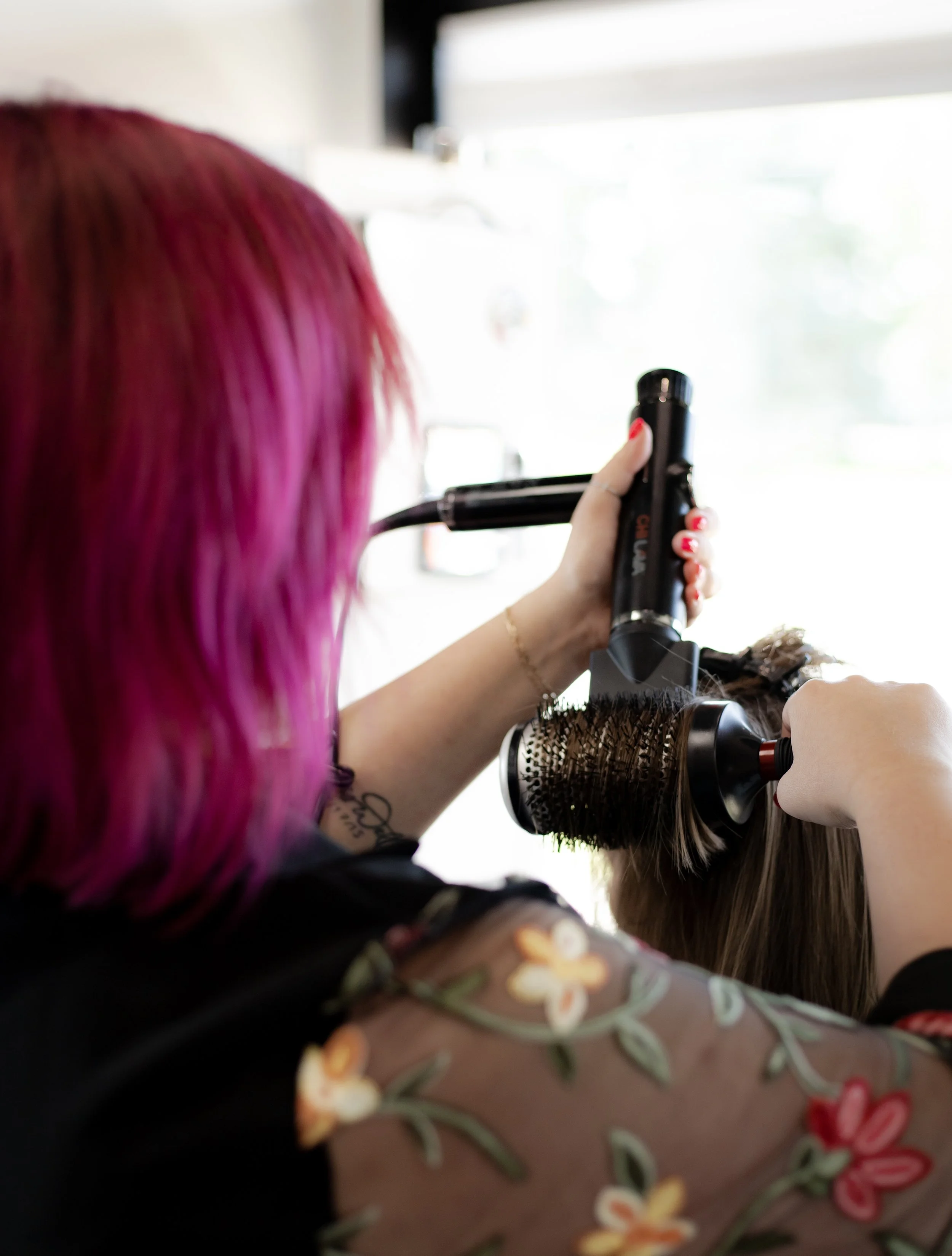 A hairstylist with pink hair using a round brush and a hairdryer to style a woman's hair.