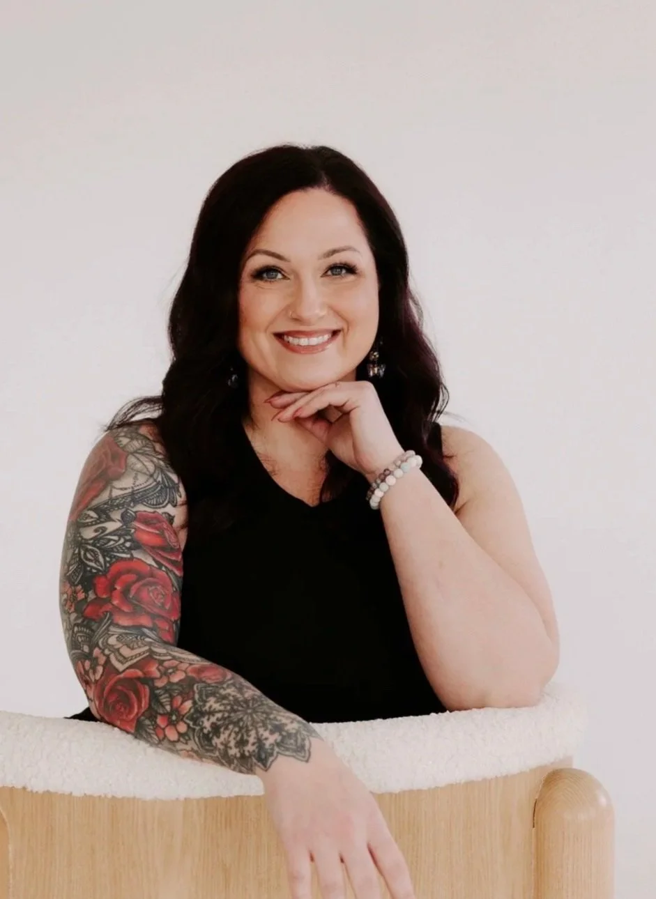 A woman with purple and black hair, tattoos on her arm, wearing a black button-up shirt, smiling against a dark background.