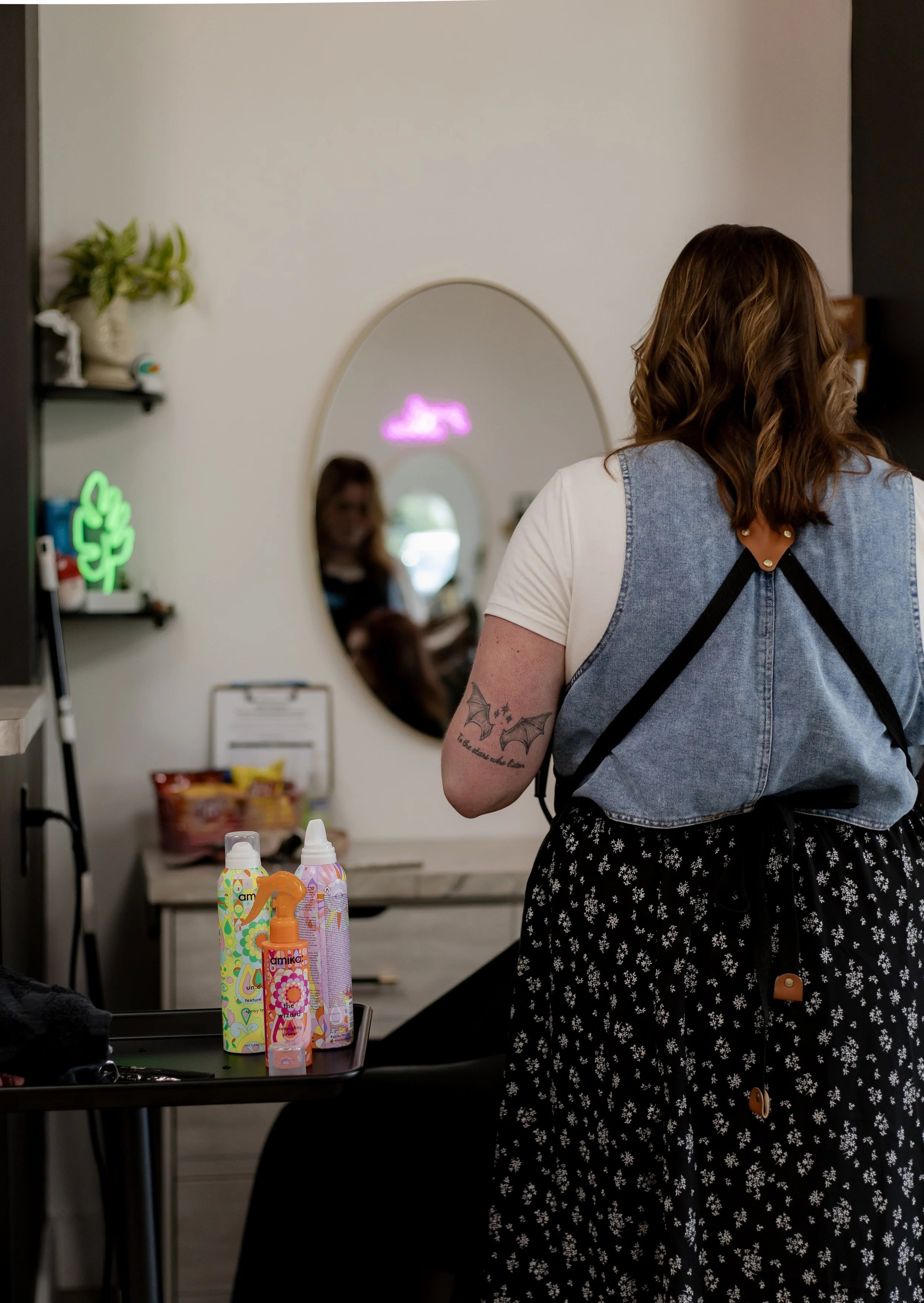 A woman with brown hair and a tattoo on her arm stands in a salon or store, facing a mirror. On her back, she wears a light blue denim vest over a white t-shirt, and a black floral skirt. On a table nearby are some colorful bottles of hair or skincar