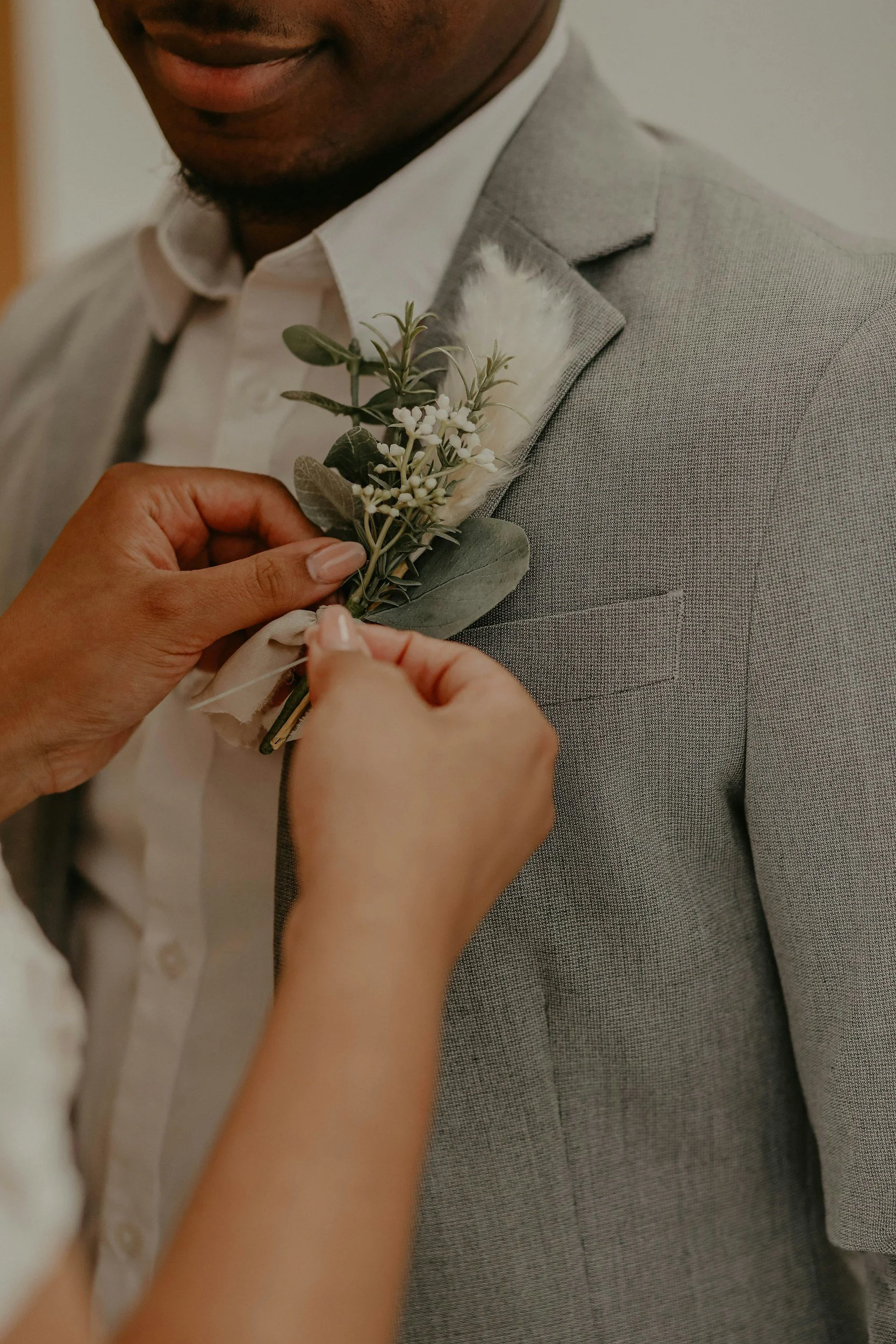 Person pinning a boutonniere with white flowers and green leaves onto a man's gray suit jacket.