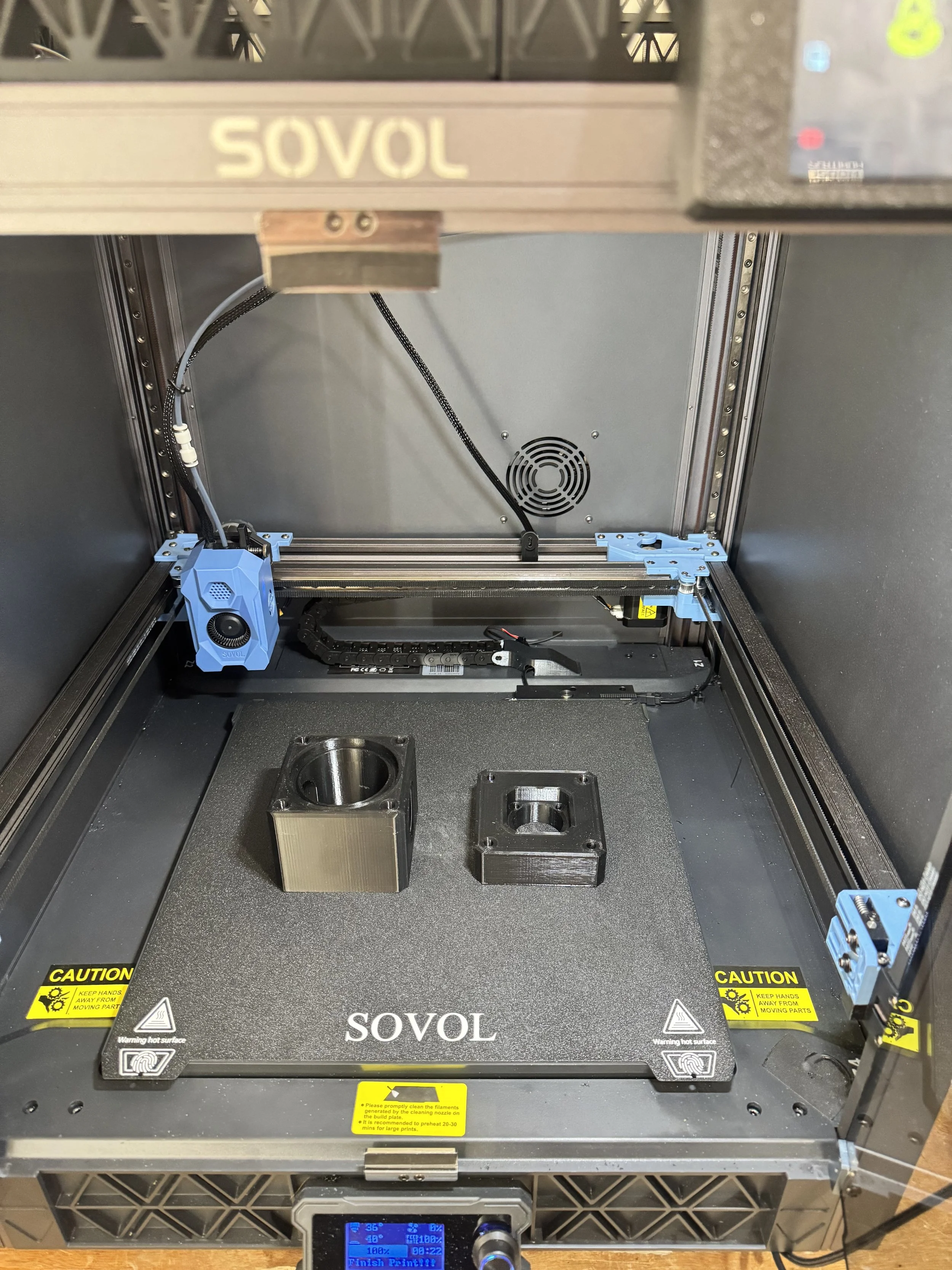 A Sovol 3D printer with two printed objects on the print bed, one cylindrical and the other square with a hole in the center, in a machine with warning labels and components.