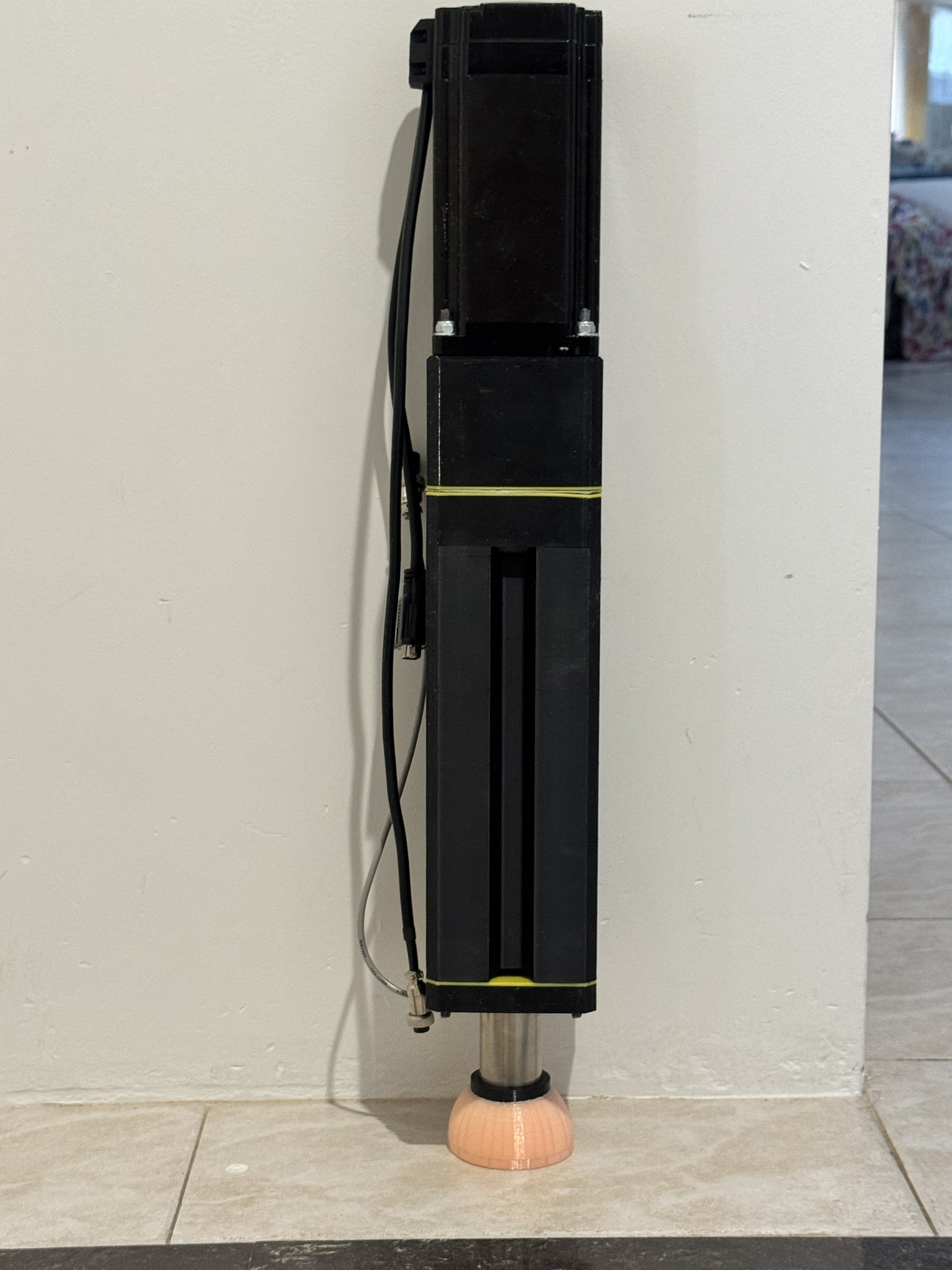 A large black industrial device, possibly a laser or scientific instrument, mounted on a stand with a copper-colored base, positioned against a wall on a tiled floor.