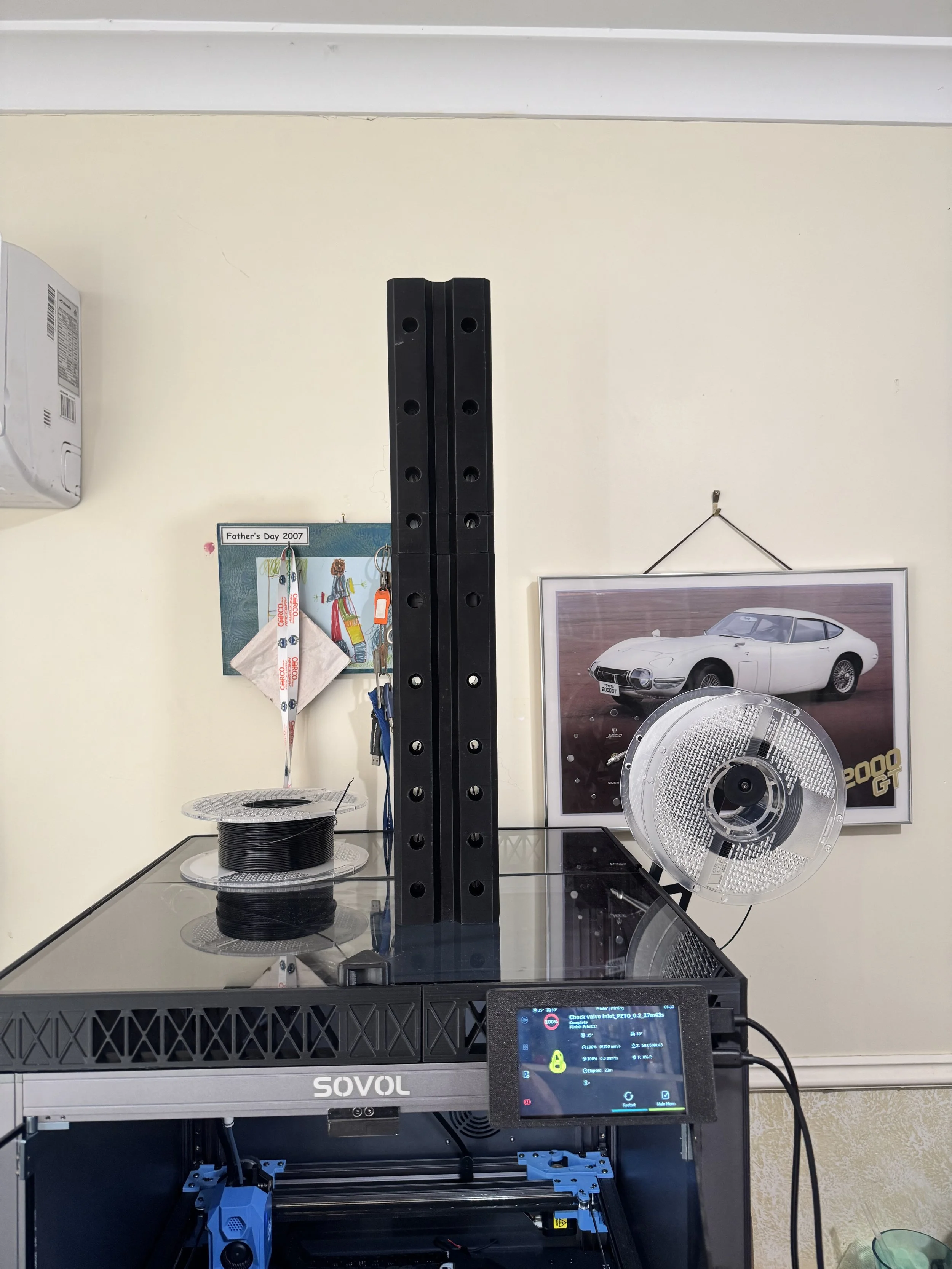 A 3D printer on a black metal frame with spool of black filament, a controlling screen, and a fan, situated in a room with a BMW poster on the wall.