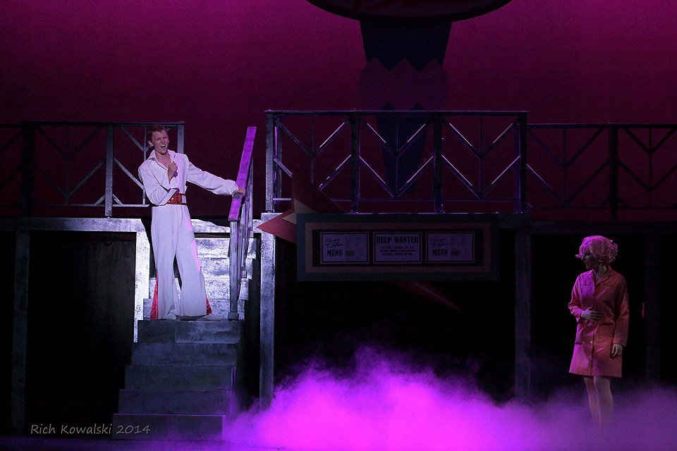 Two actors on a stage portraying a scene with stairs, purple fog, and dim lighting. One actor stands on the stairs dressed in white with a microphone, and the other stands below dressed in pink. Grease Teen Angel Count Basie Theatre