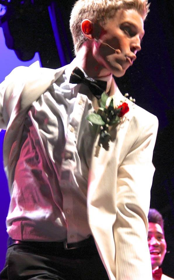 A young man dancing in a white tuxedo with a black bow tie and a red flower boutonniere, performing on stage with a microphone attached to his face, and another person smiling in the background. Footloose Ren Berklee College of Music