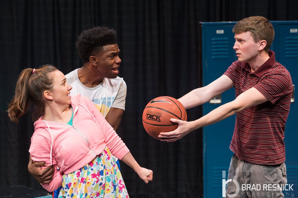 the male on the right, wearing a striped red polo shirt, is holding out a basketball toward the female, who looks scared, while the other male, with curly hair. Children's theater educational New Kid George Street Playhouse