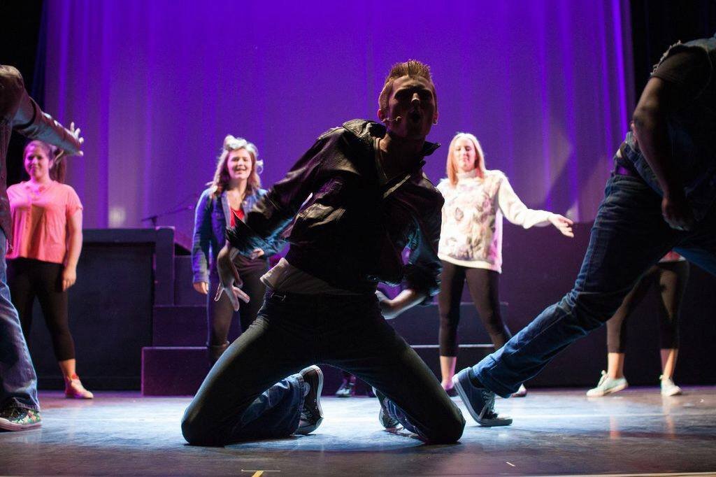 A man kneeling on stage with his mouth open, surrounded by women standing and moving in the background during a theater or dance performance. Footloose Ren Berklee College of Music