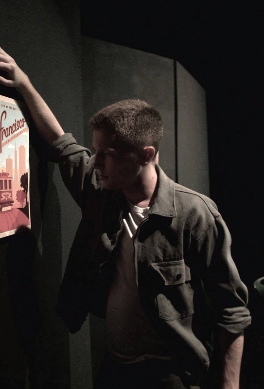 Young man with short hair wearing a gray jacket standing in a dark room, leaning against a wall, with a poster on the wall beside him. Dogfight Boland Harvard