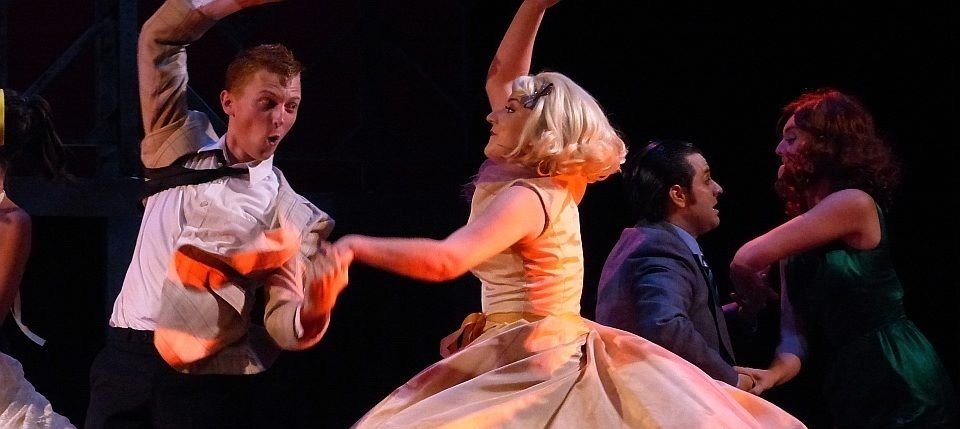 Four performers in vintage clothing dancing on stage, with a woman in a beige and yellow dress spinning and a man and a woman in the background watching. Grease Doody Count Basie Theatre