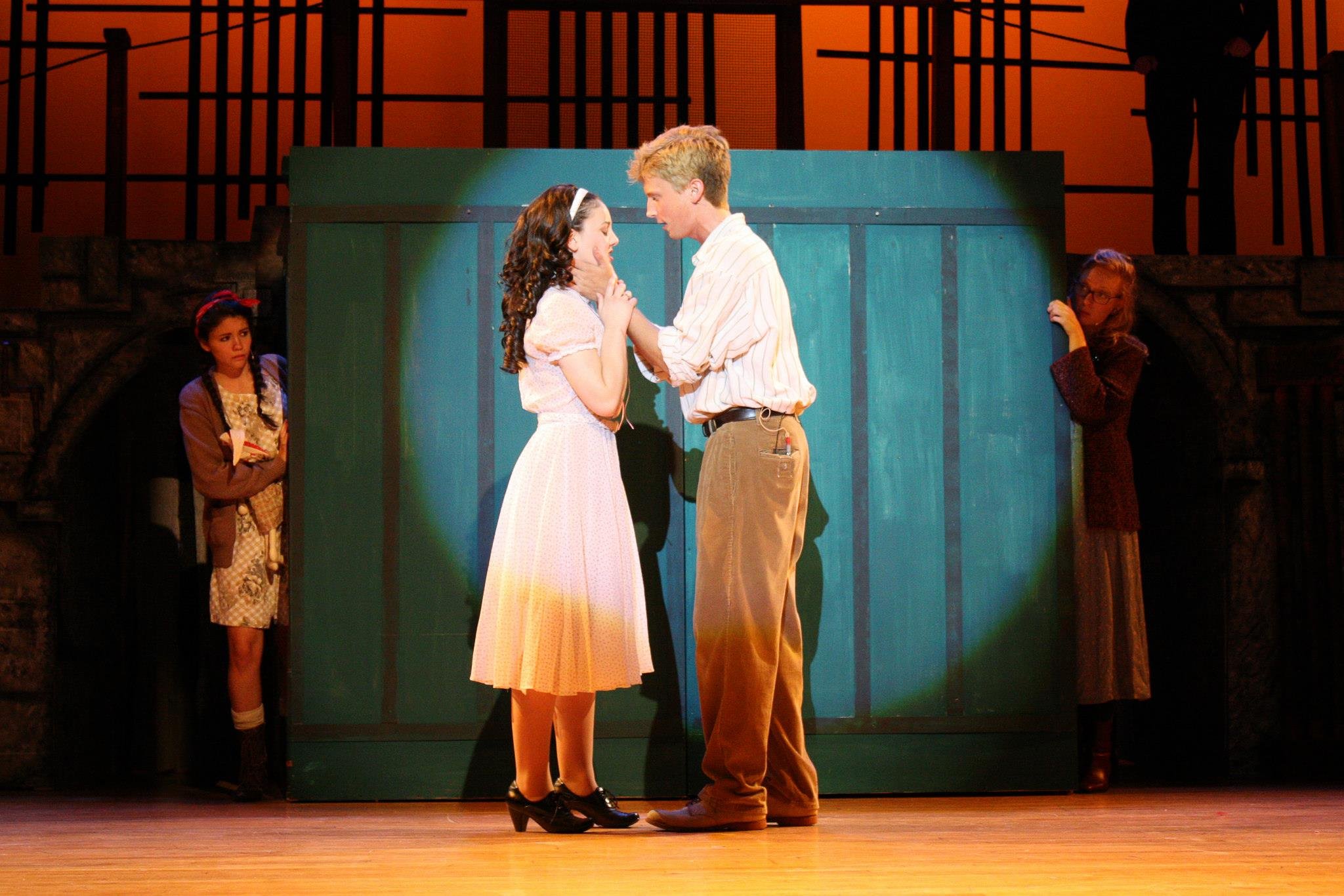 A scene from a theatrical play with two actors in the center, a young woman in a white dress and a young man in a striped shirt and pants, touching tenderly. Urinetown Bobby Strong