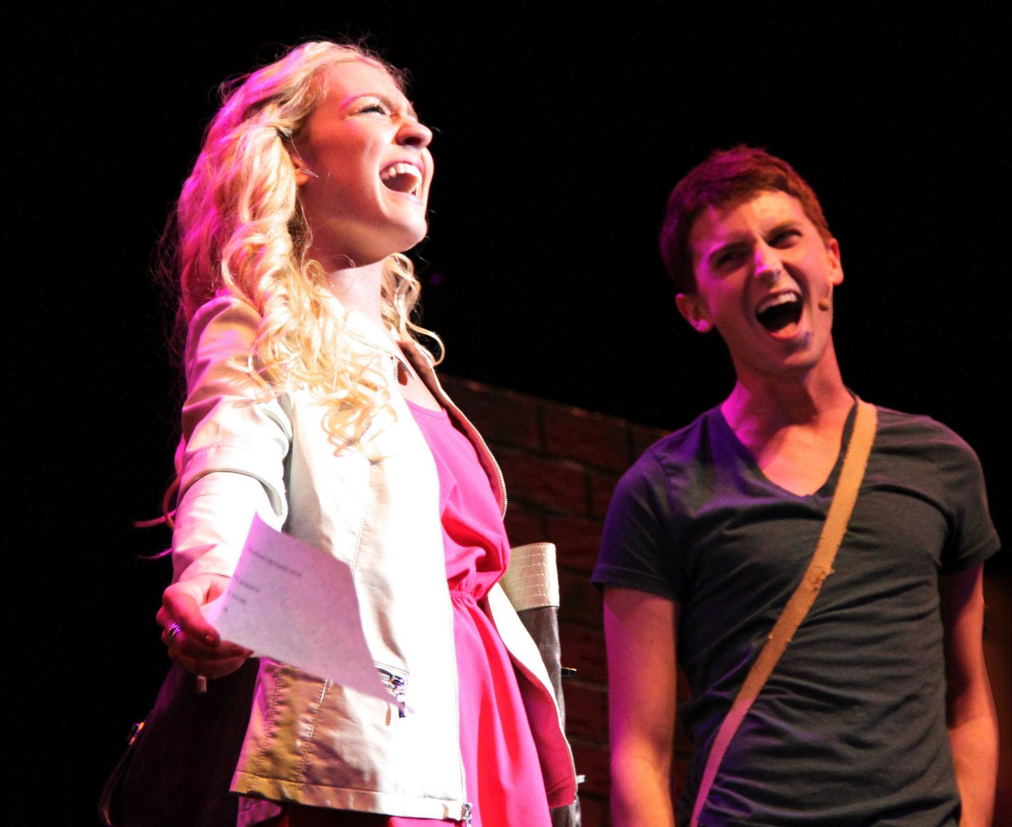 Two people on stage, singing passionately, under pink and purple stage lighting. The woman on the left has curly blonde hair and is wearing a pink dress with a white jacket. Legally Blonde Emmett Berklee College of Music.
