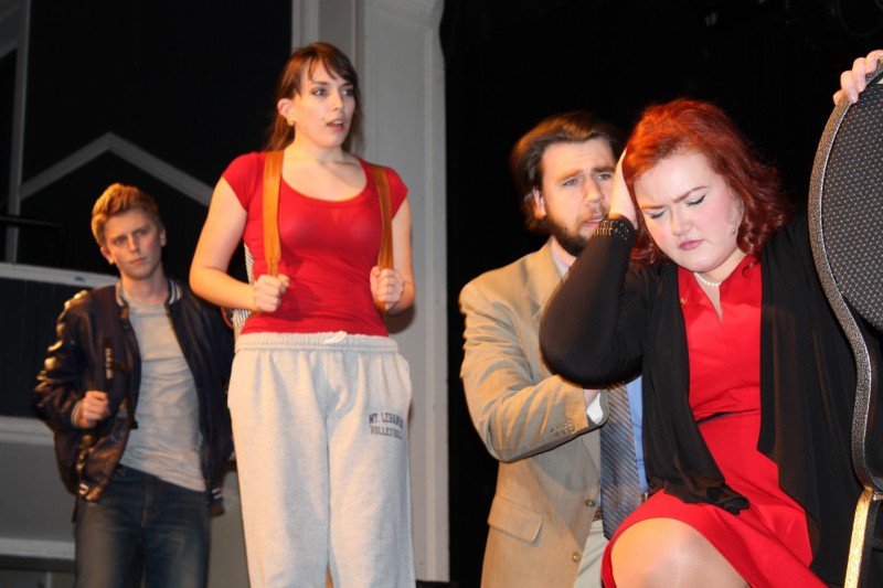 Four young adults on a stage; A woman with brown hair in a red shirt and grey sweatpants stands nearby, and another young man in a leather jacket looks on in the background. Next to Normal Gabe Berklee College of Music