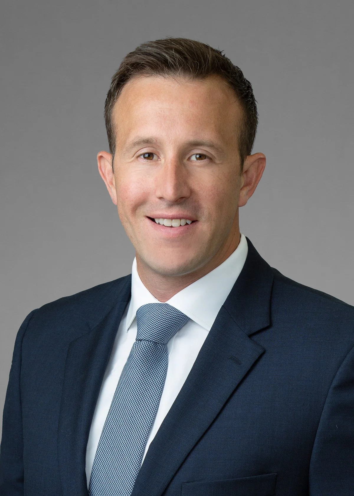 Headshot of a smiling Caucasian man with short brown hair, dressed in a dark navy suit, white shirt, and a light blue tie, against a gray background.