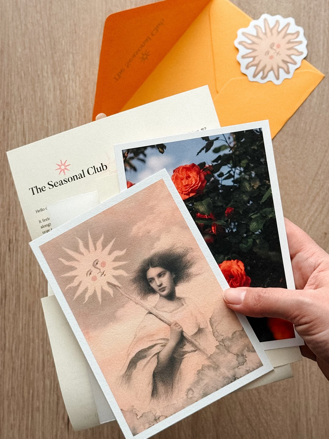 The Seasonal Club Beltane edition, held in hand: a peach-toned art print of a young woman holding a staff with a smiling sun on top, a photo of red roses, a letter, and an envelope with a sun sticker.