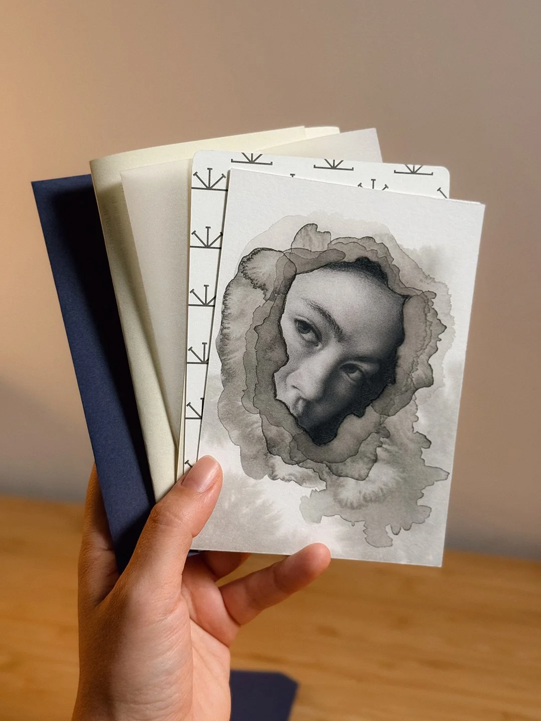 The Imbolc snail mail edition, fanned out in hand: a graphite portrait of a woman's face showing through layered grey ink blooms, a patterned card, cream stationery and a navy envelope.