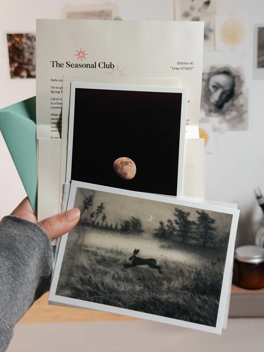 The Seasonal Club spring edition, "Leap of Faith": a charcoal-toned print of a hare leaping through a misty meadow under a crescent moon, a photo print of the moon, a letter and a mint-green envelope.