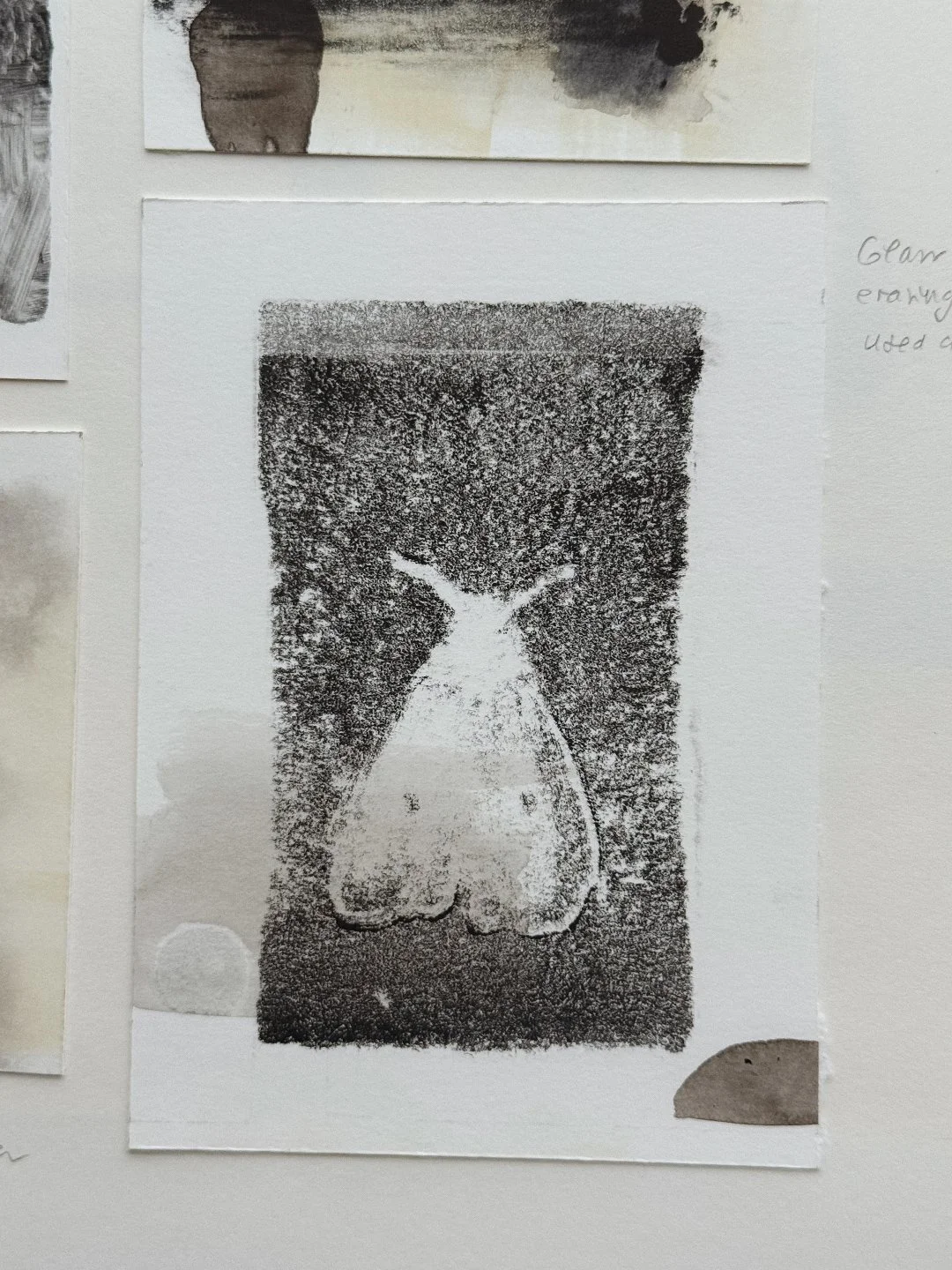Small monotype of a pale moth silhouette lifted out of a dark, grainy field of ink, mounted on a sketchbook page next to other ink experiments.