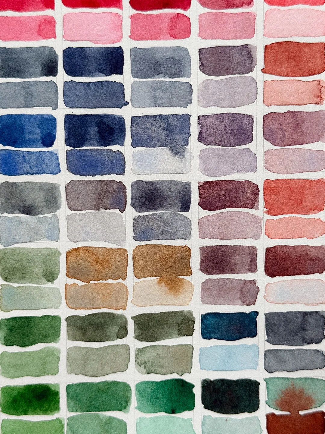 Detail of watercolour mixing swatches in muted greys, dusty violets, olive greens and granulating copper browns. A Phthalo Green swatch has a rust-red bloom spreading across it.