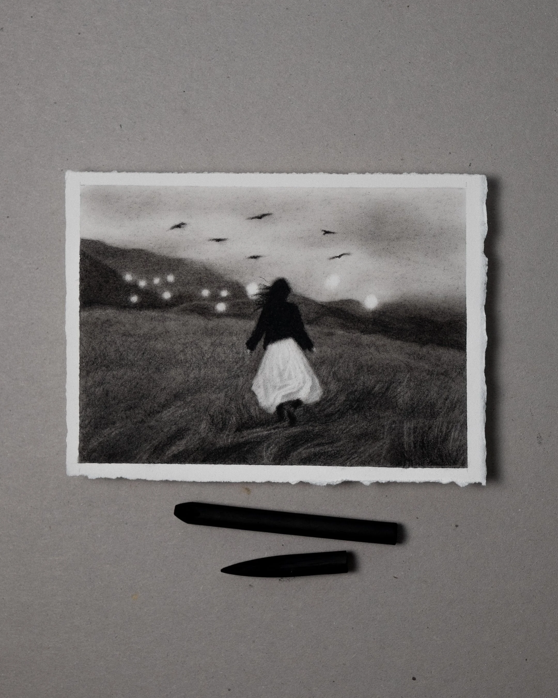 A black-and-white graphite and charcoal drawing of a woman running through an open field, towards the forest in the background, birds flying in the sky, and lights or orbs scattered across the scene.