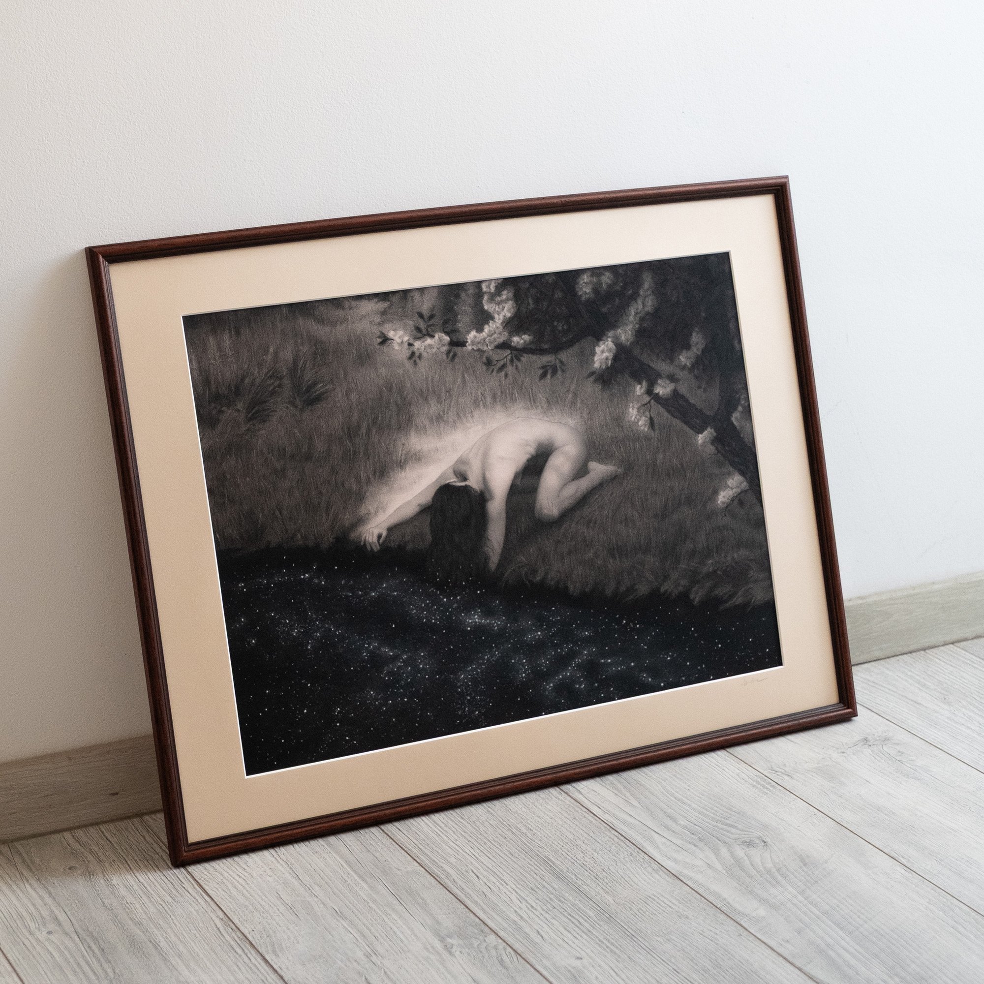 The drawing "Surrender" by avidoma depicting a woman kneeling on a river bank, framed, leaning against a white wall.