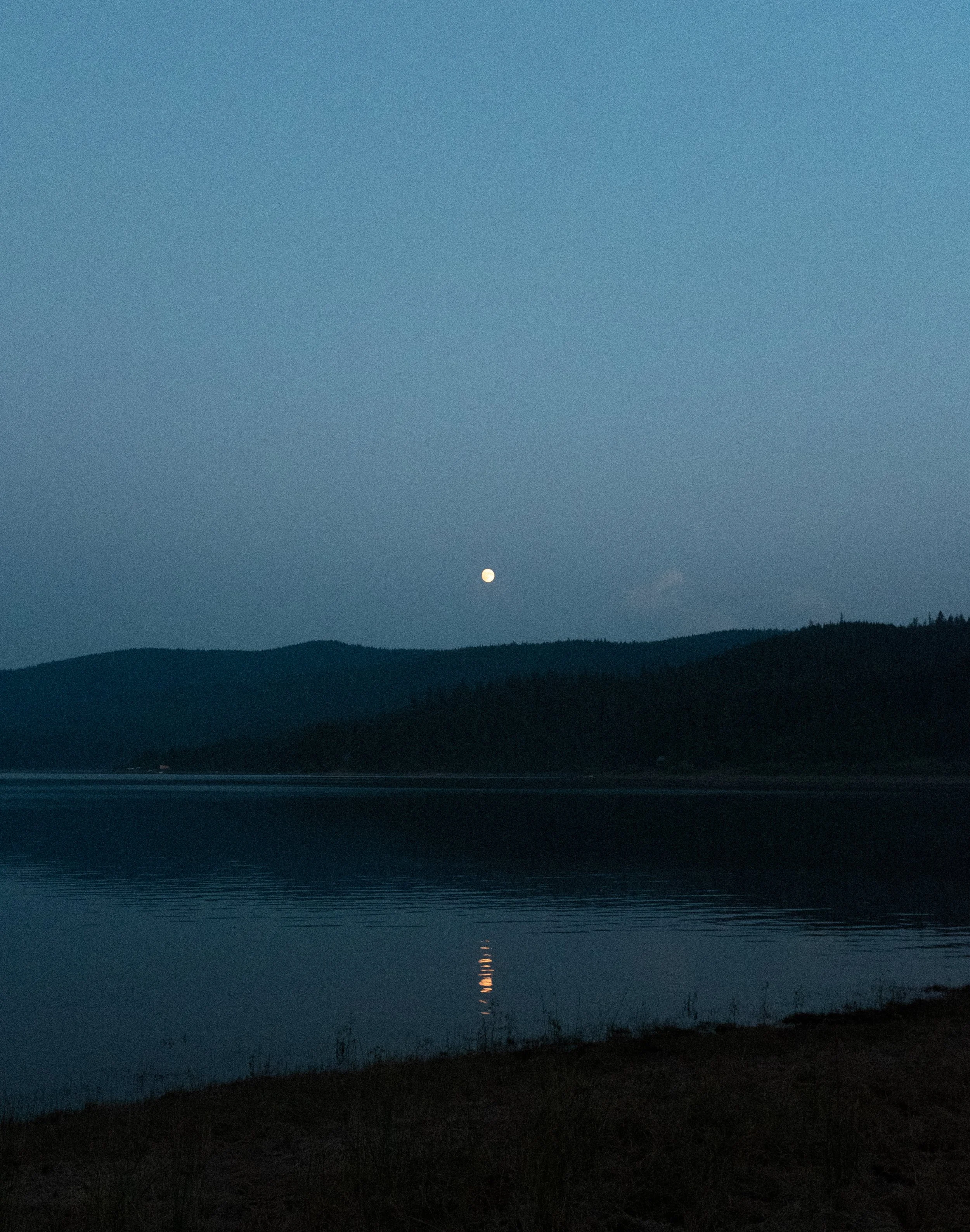 A serene evening landscape featuring a calm lake with the reflection of a bright moon, surrounded by silhouetted hills and a clear blue sky.