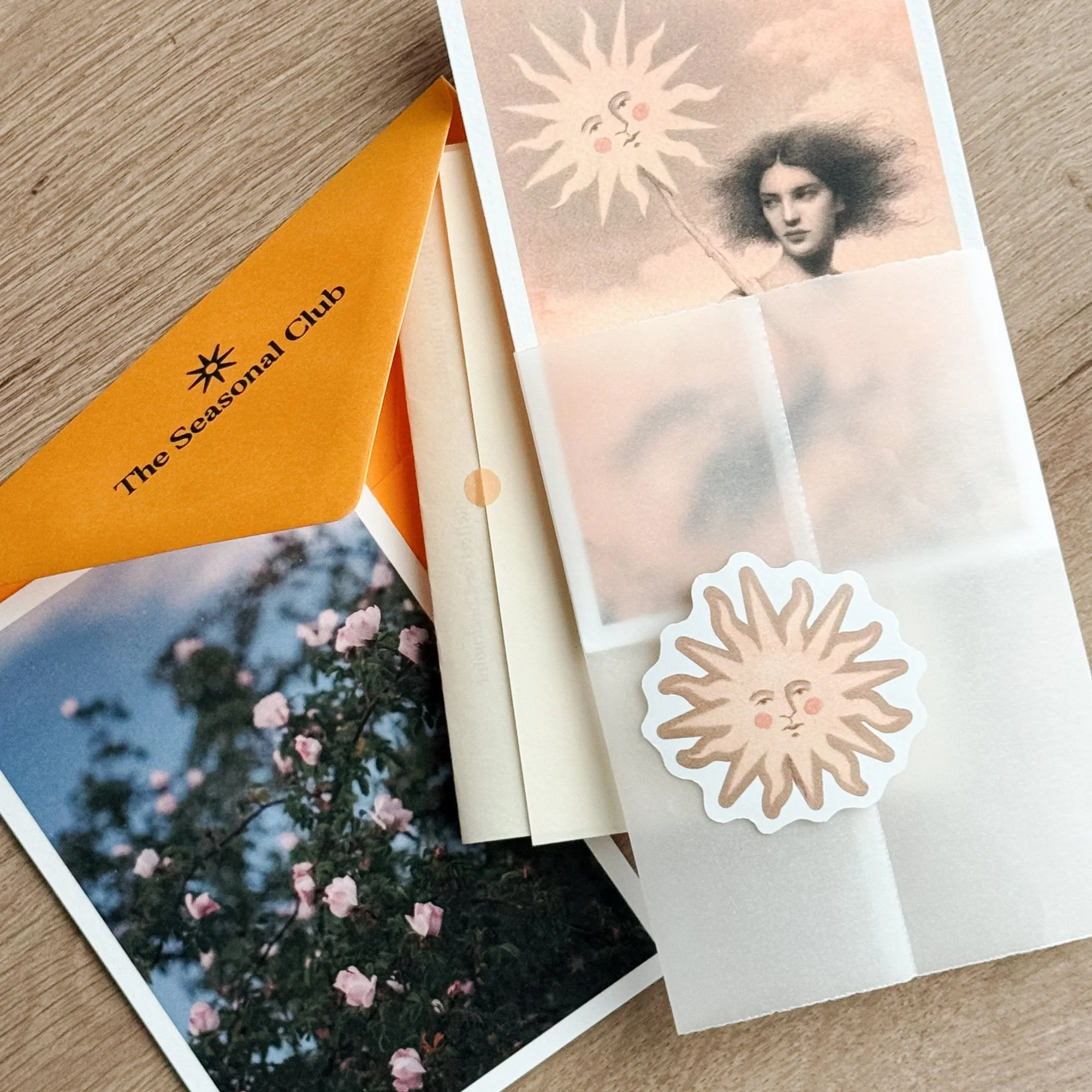 Flat lay of an art snail mail yellow envelope, with a matching floral postcard, surreal art print of a woman, and an alchemy style sun sticker