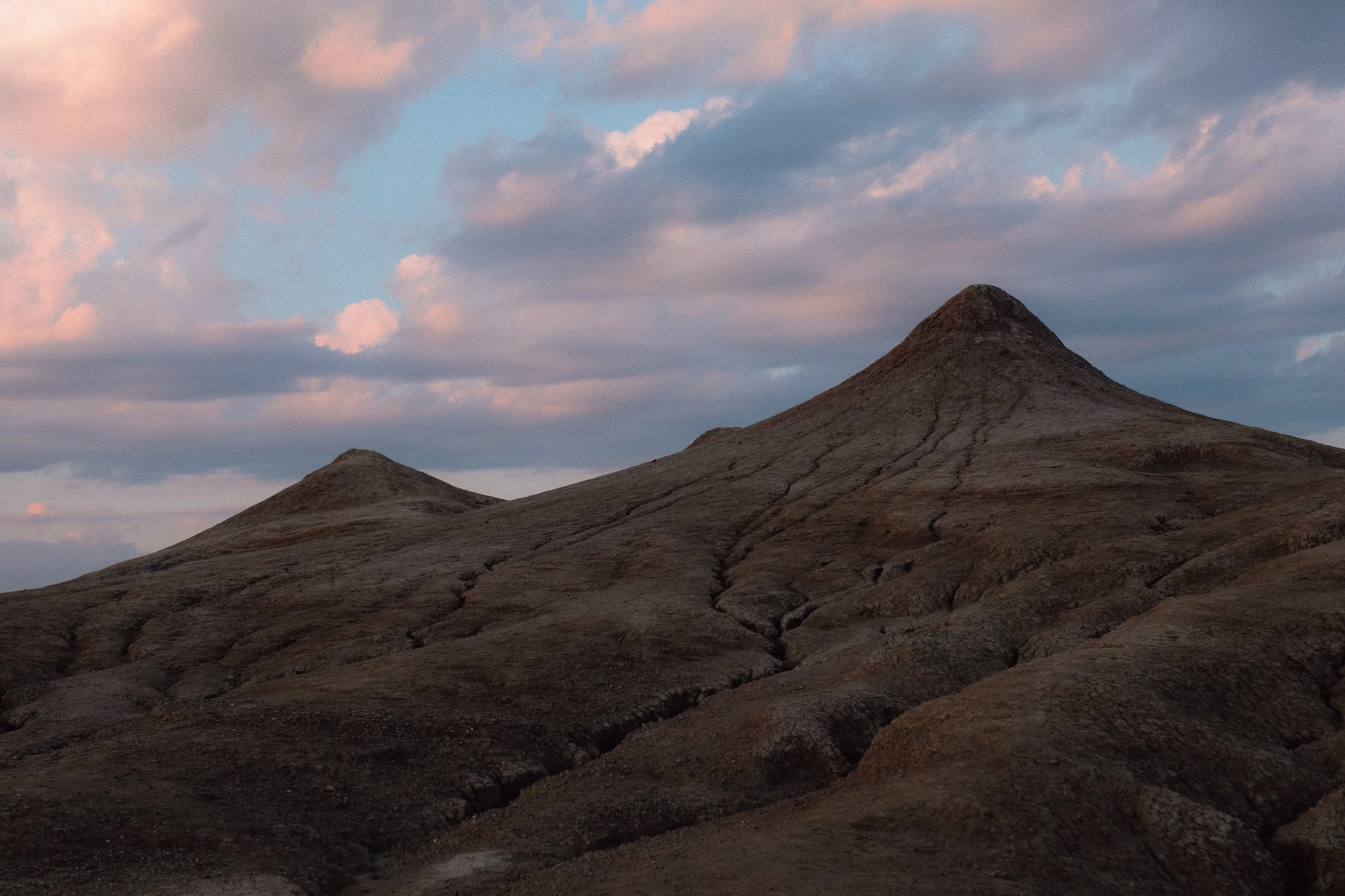 A landscape featuring two brown, rounded hills at Vulcanii Noroiosi under a sky with scattered pink and blue clouds at sunset.