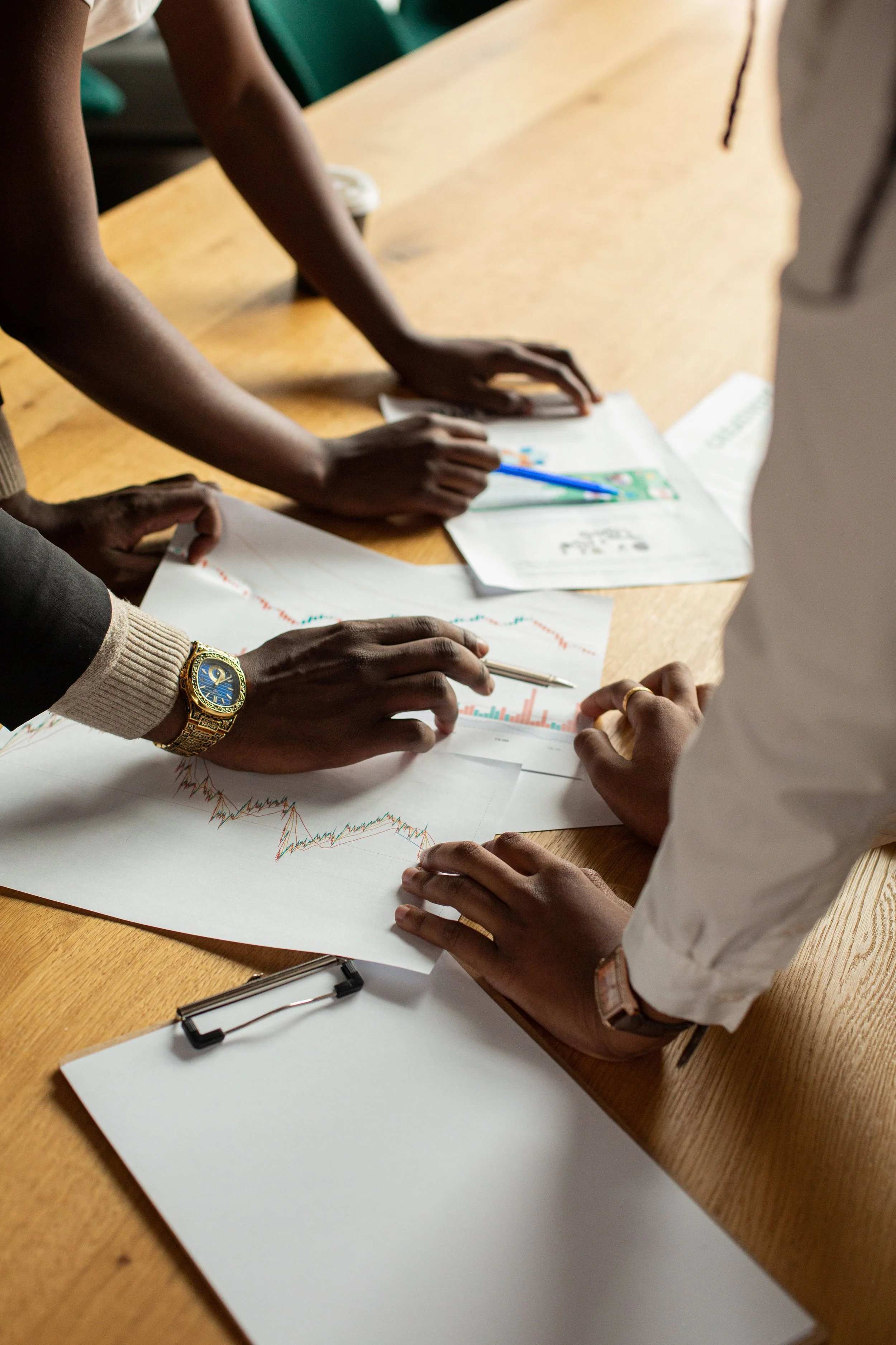 Multiple people gathered around a wooden table looking at financial charts and graphs on sheets of paper.