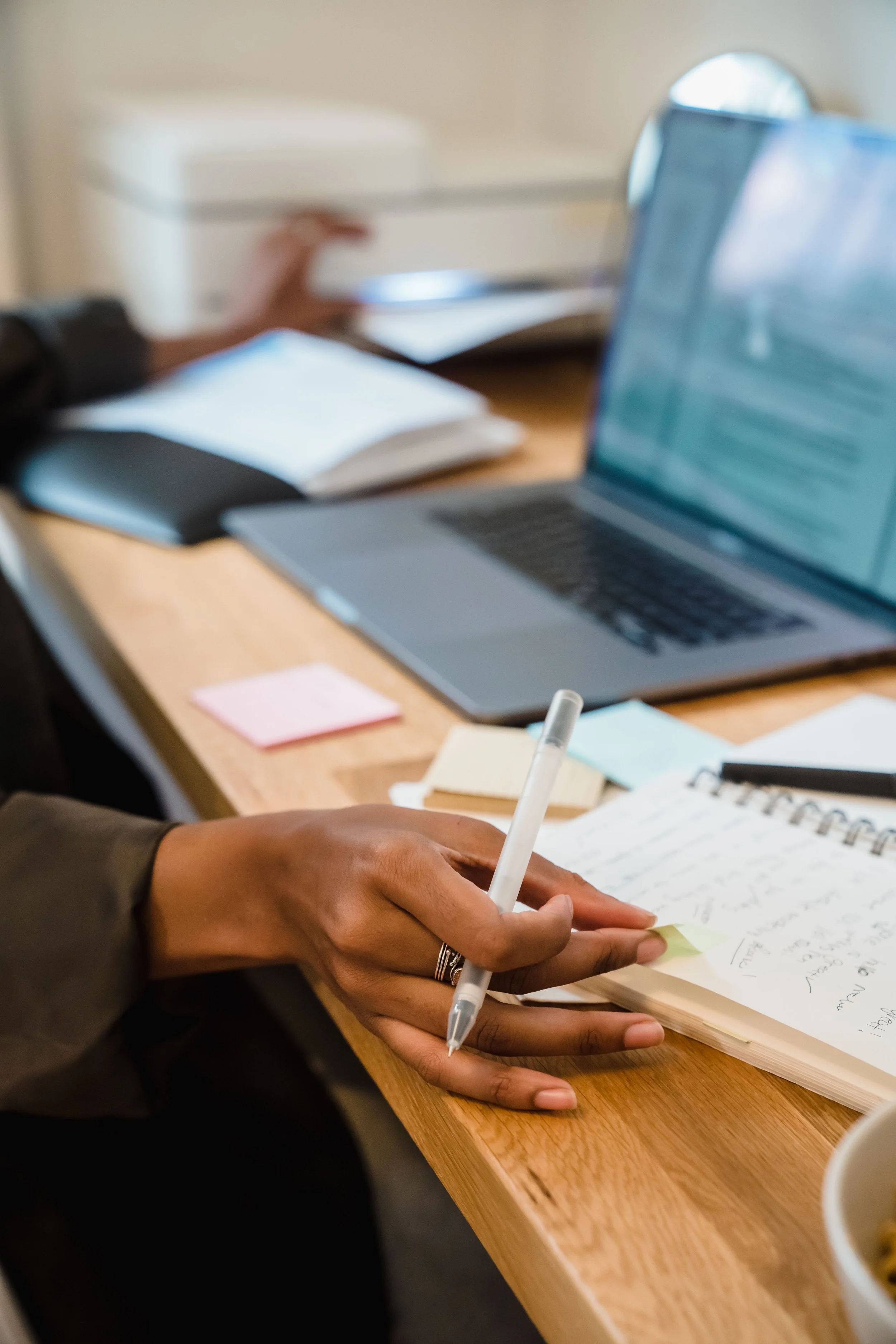 A person in a dark jacket is writing in a notebook with a white pen on a wooden desk. There is a laptop, a pink sticky note, and some papers on the desk.