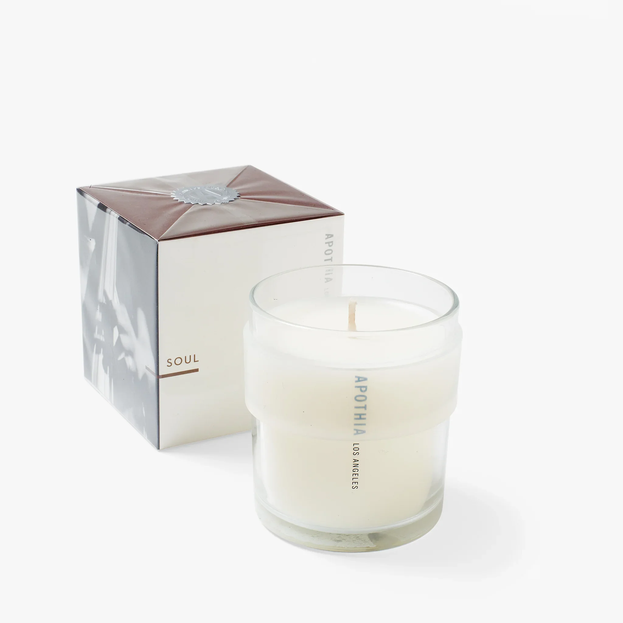 White scented candle in a glass container with black text, next to a metallic box with white and brown packaging, labeled 'SOUL' and 'APOTHEA LOS ANGELES'.